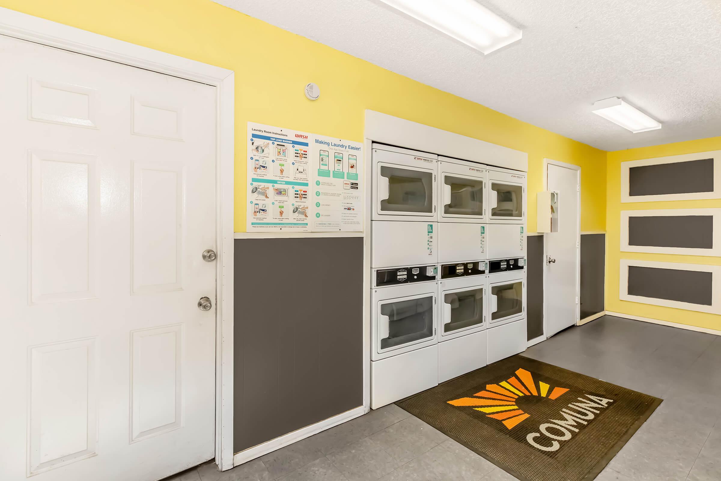 A bright laundry room featuring yellow walls and gray accents. On one side, there are several stacked washer and dryer units. A door leads outside, and a welcome mat at the entrance reads "COMMUNITY." A poster on the wall provides laundry instructions and guidelines.