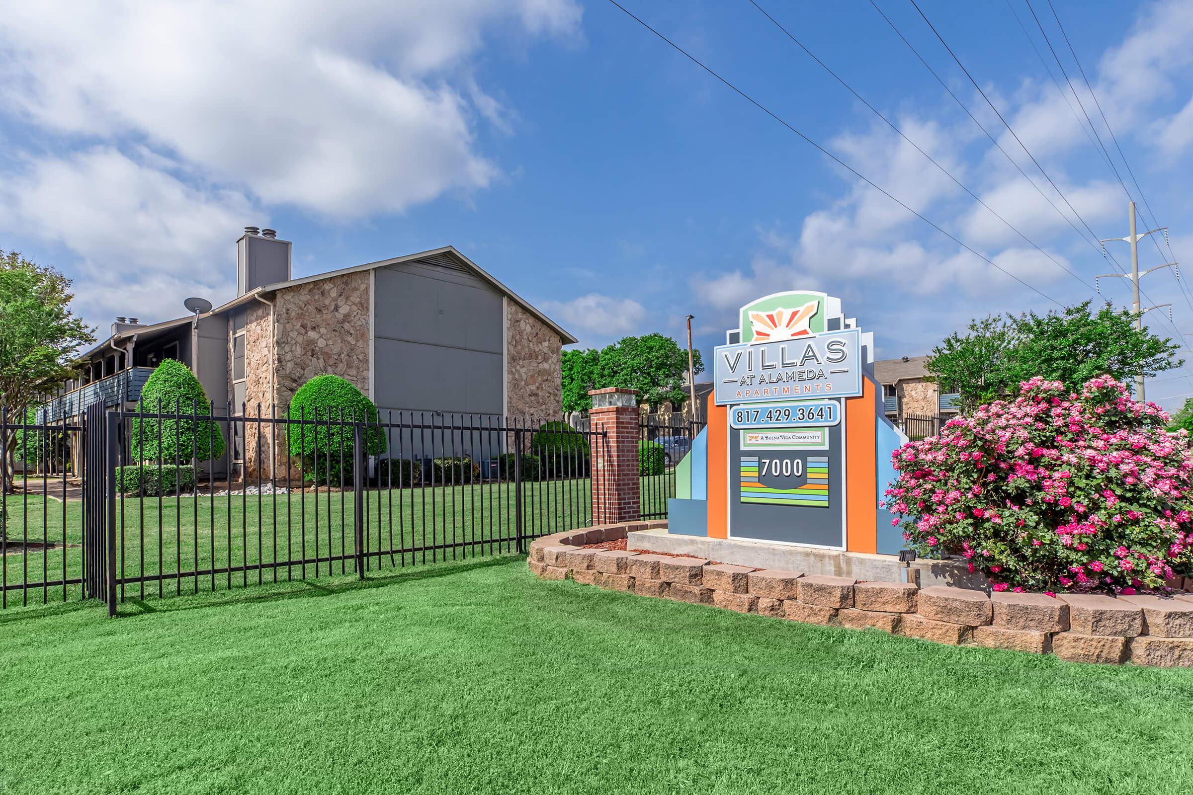 A stone and stucco apartment complex with a landscaped front yard featuring manicured bushes and flowers. A prominent sign reads "Villas at Alameda," indicating the name of the property, with contact details below. The scene is bright and well-maintained, with a clear blue sky.