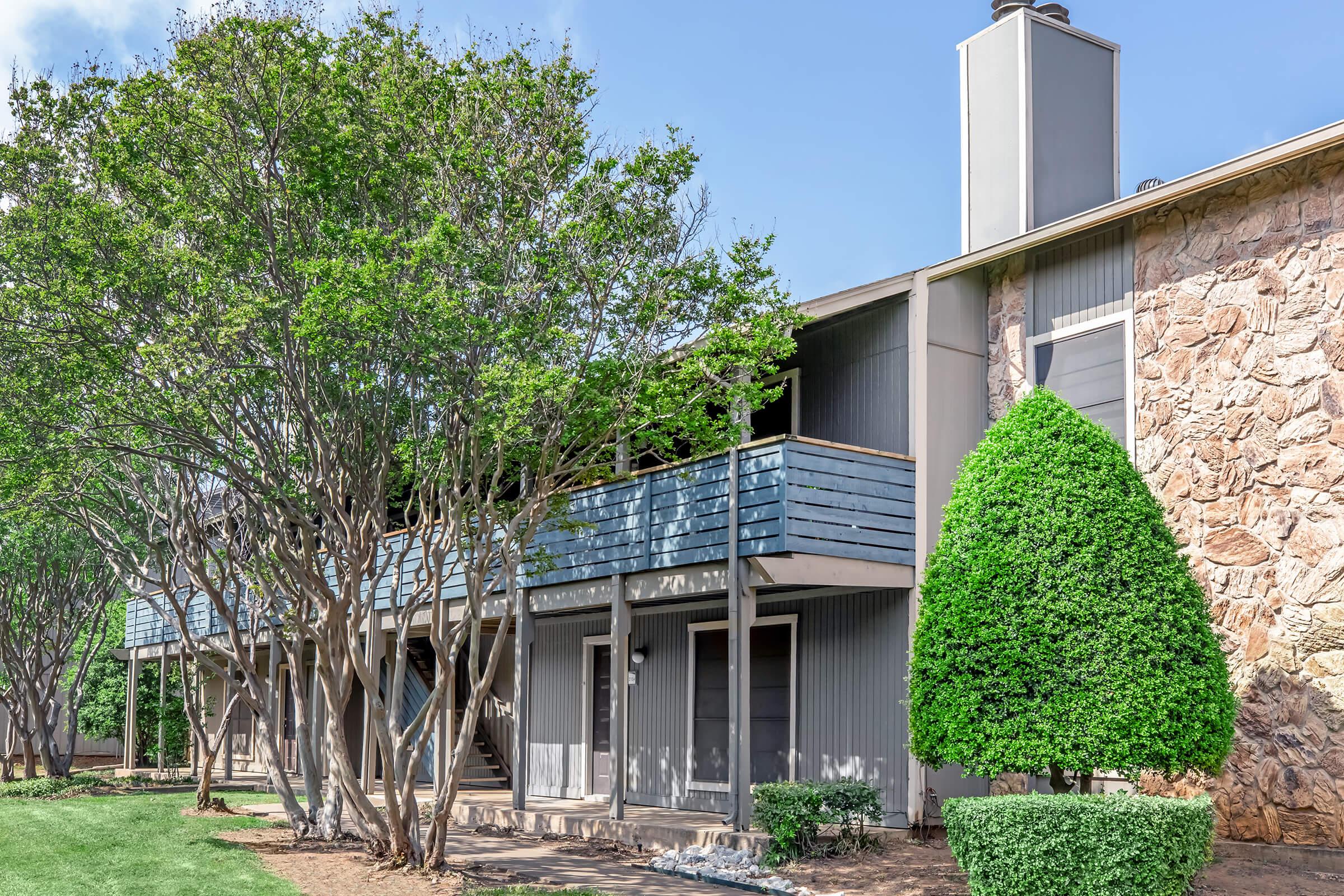 A modern two-story residential building with a combination of stone and siding exteriors. It features green trees and well-manicured landscaping, along with balconies on the upper level. The setting is bright and inviting, suggesting a well-maintained apartment complex.