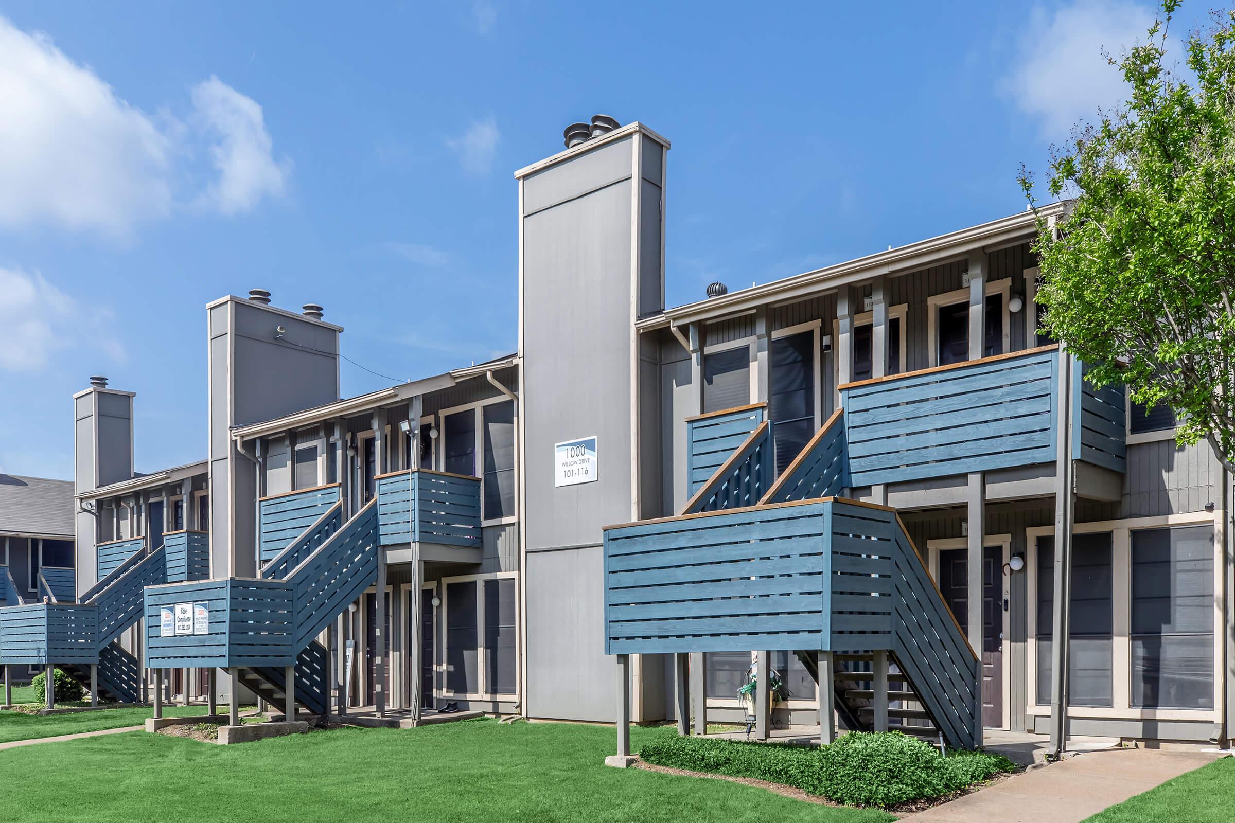 Two-story apartment building with a modern design, featuring blue balconied staircases and a grassy lawn. The exterior has light gray siding, with a clear blue sky above.