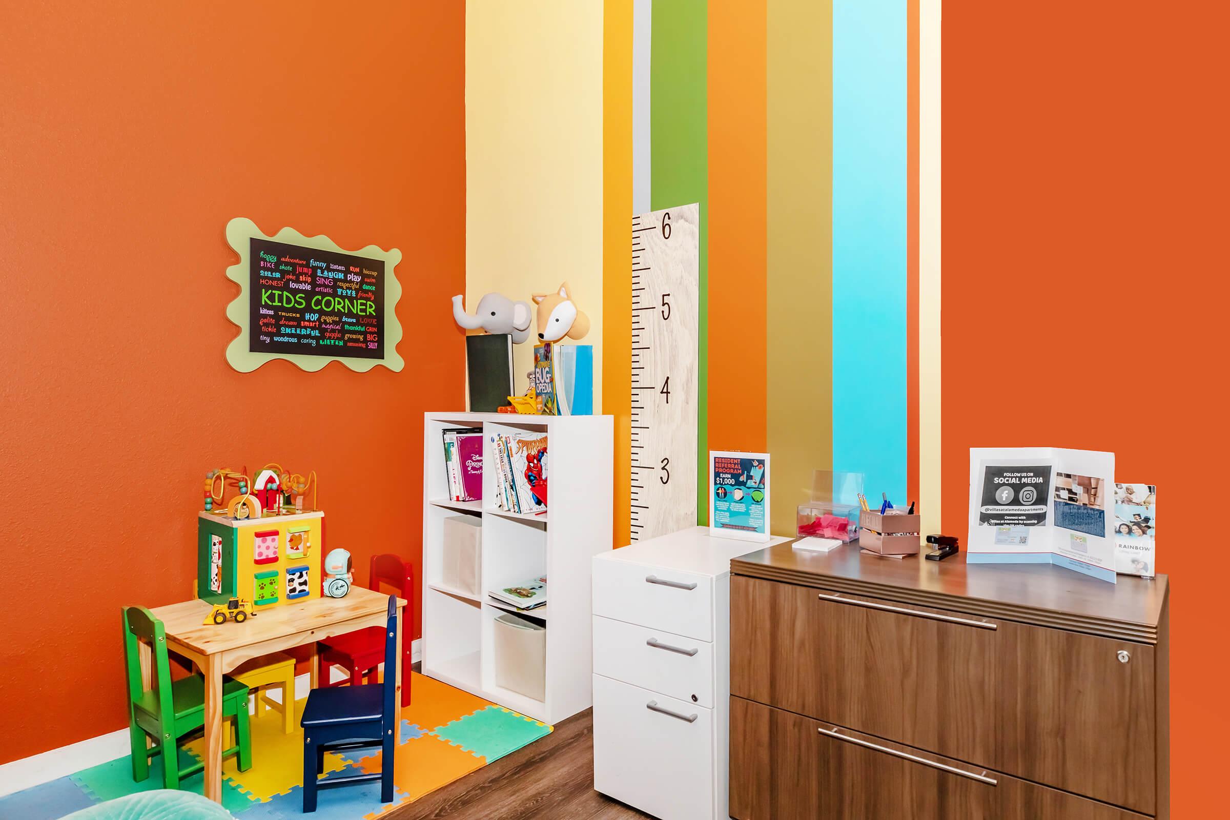 A colorful children's corner featuring a small table and chairs, various toys on a play mat, a tall ruler against the wall, a white storage unit with books, and a wooden cabinet. The walls are painted in vibrant colors with striped patterns, creating a cheerful atmosphere for kids.