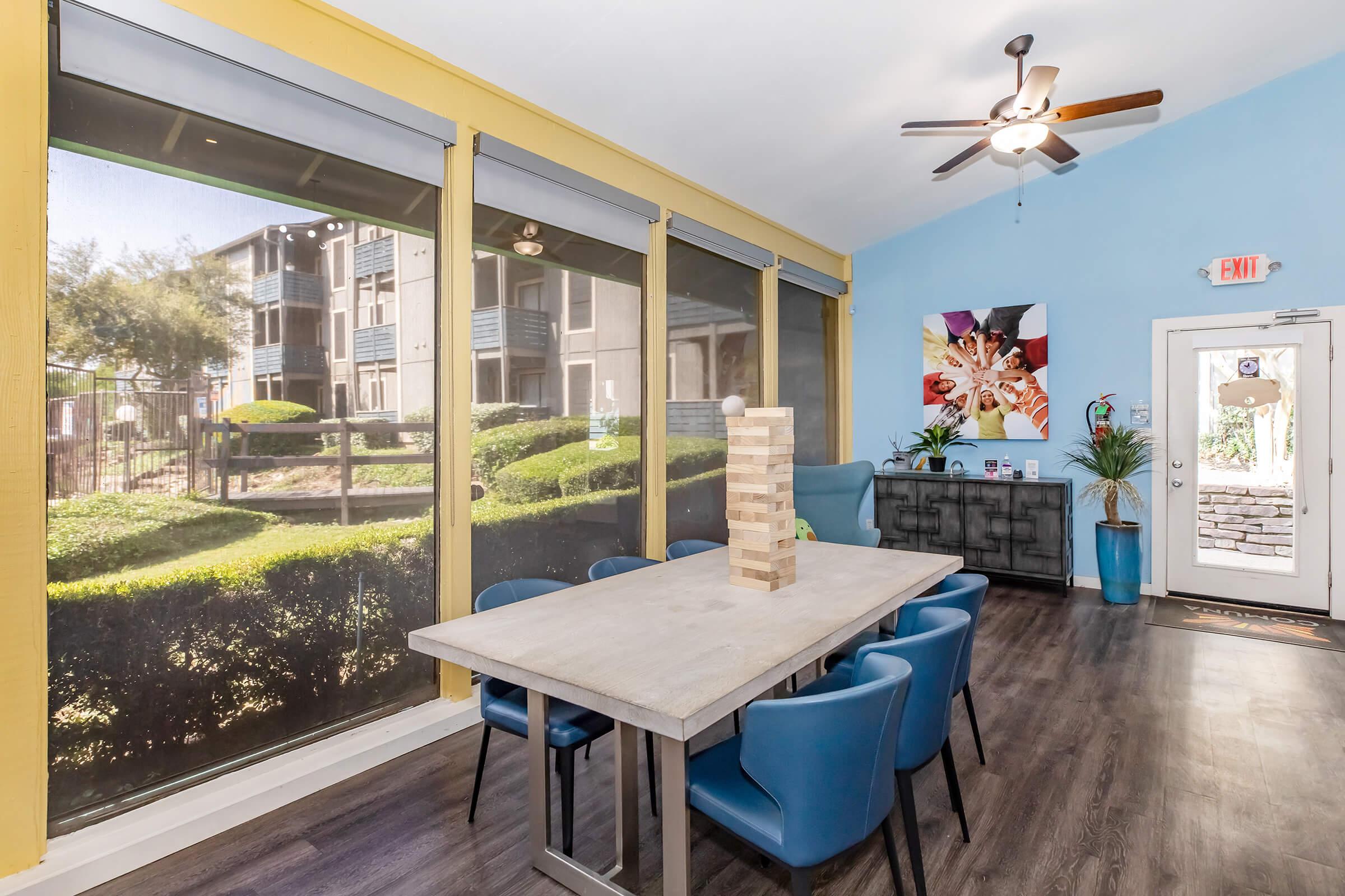 A bright common area featuring a wooden table surrounded by blue chairs, with large windows overlooking greenery and an apartment complex. A ceiling fan is visible above, and there are decorative plants and artwork on the walls, creating a welcoming atmosphere.