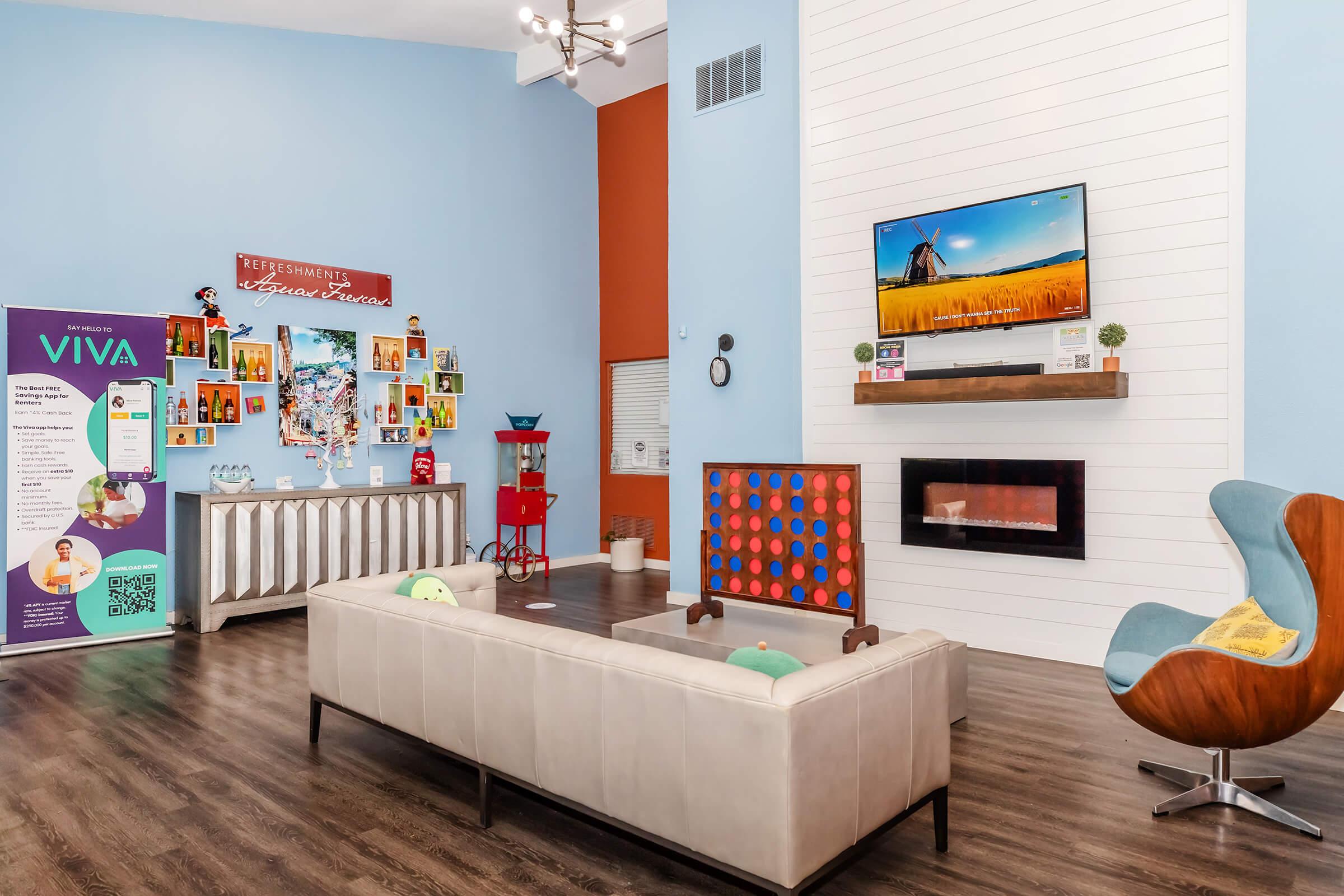 Bright and inviting lounge area featuring a modern sofa, colorful art pieces on the wall, a television displaying a landscape, and a stylish electric fireplace. The room includes a vibrant accent wall, a refreshment station with promotional materials, and contemporary seating arrangements.