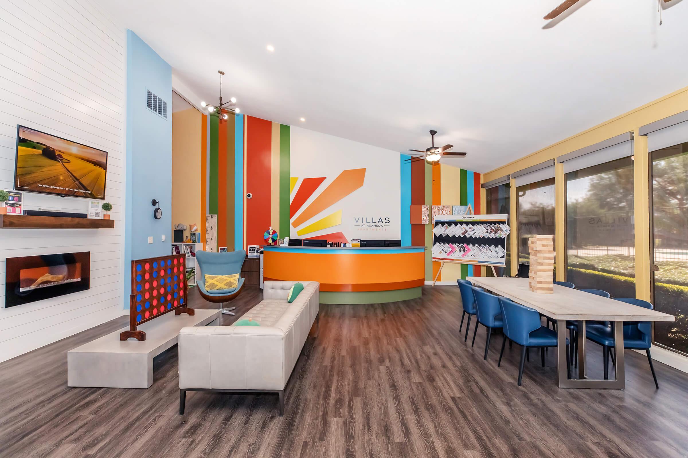 A modern community center interior featuring colorful accent walls in orange, blue, and yellow. There's a reception desk with the "Villas" logo, comfortable seating arrangements, and a large table. Large windows allow natural light, and decorative elements add a welcoming atmosphere.