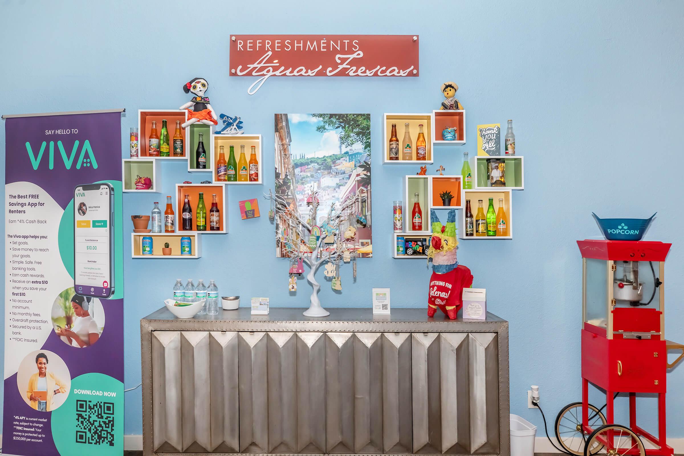 Colorful wall display featuring shelves with various drinks and decorative items, a large framed photo of a vibrant city scene, and a cart for refreshments. There's a sign reading "Refreshments Aguas Frescas" and a promotional banner for a service called VIVA on the left.