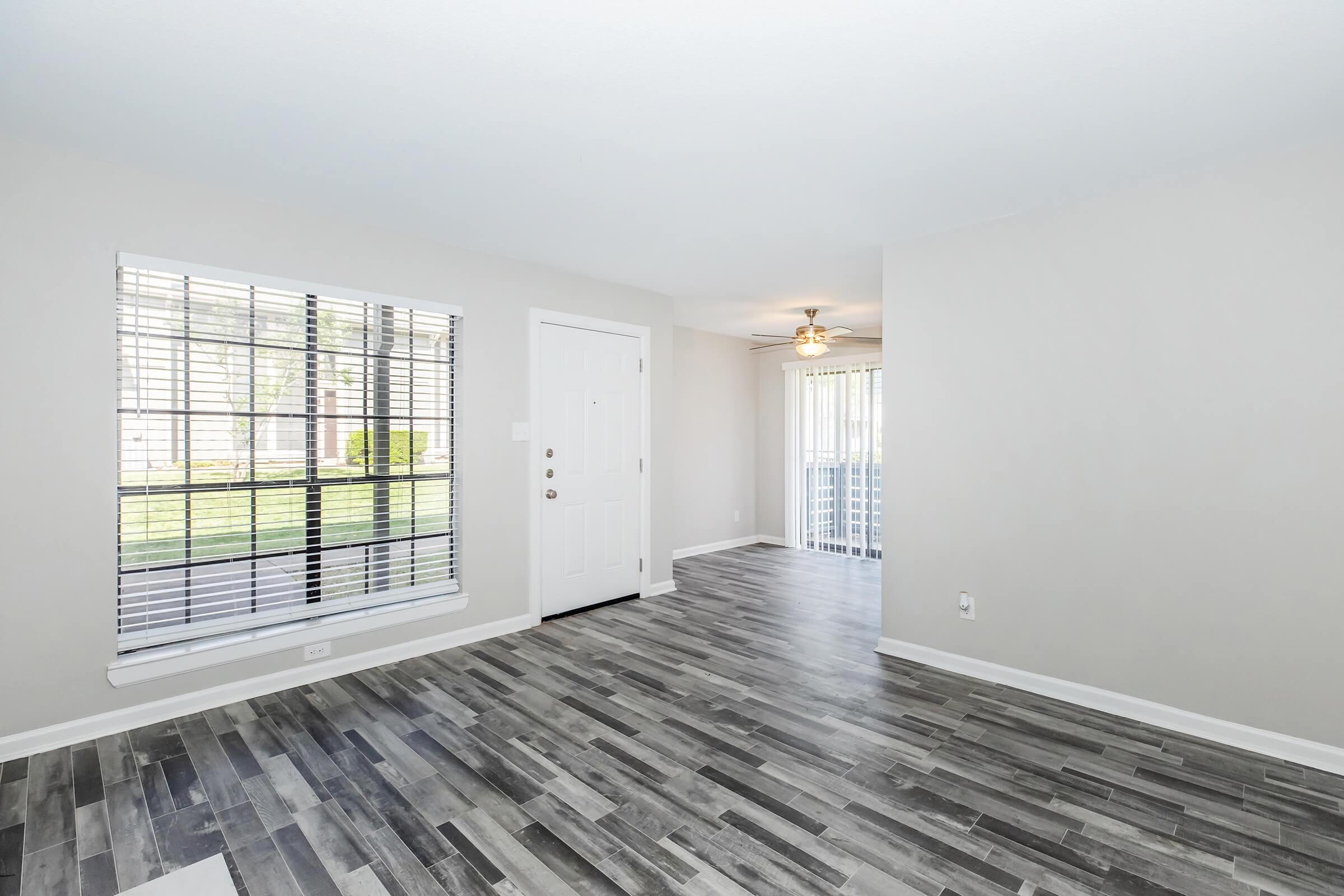 Spacious interior of a living room featuring a white door, large window with blinds, and light gray walls. The floor is adorned with wood-like tiles, and there’s an adjacent area with a ceiling fan visible. Natural light pours in from the window, creating an inviting atmosphere.
