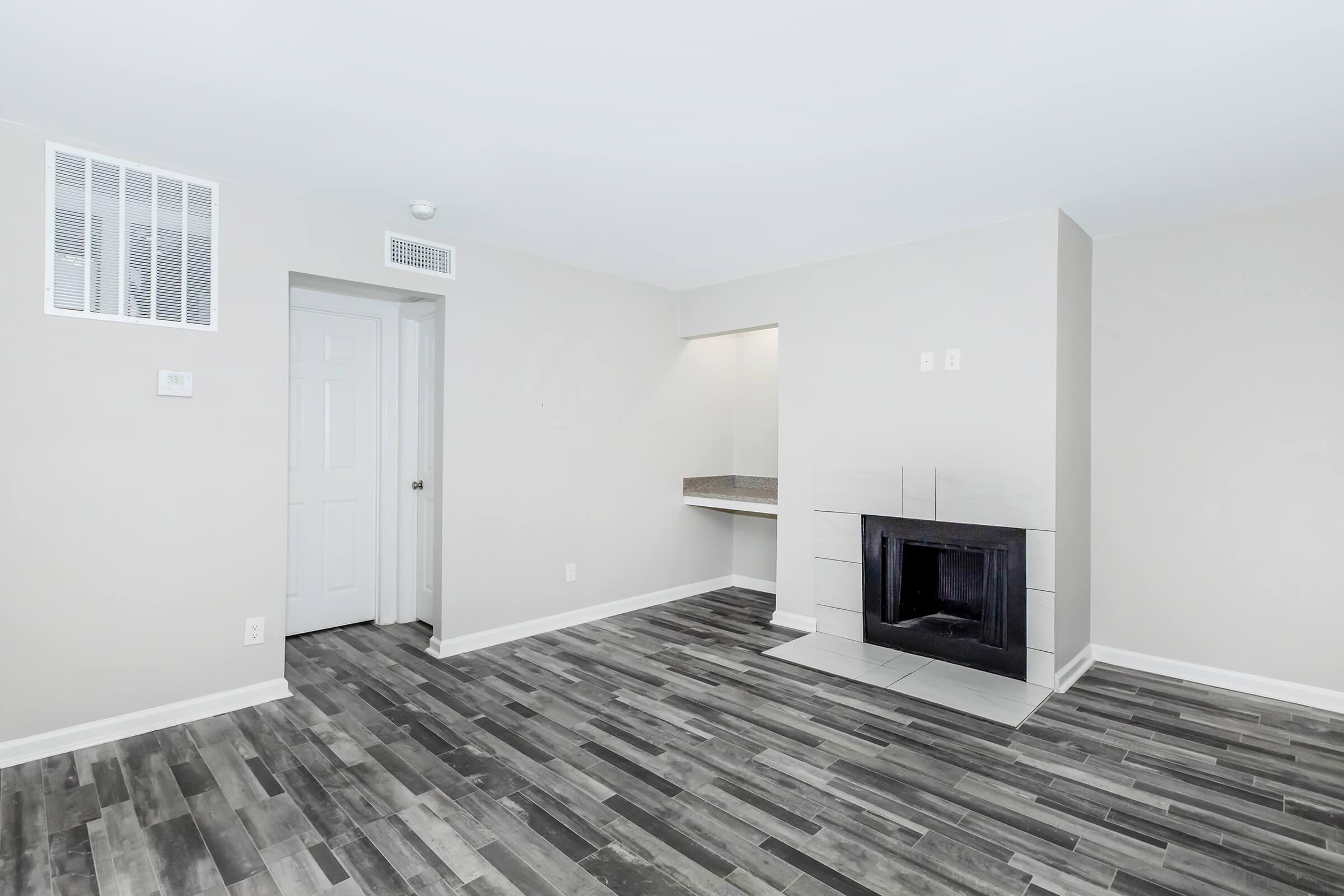 A spacious, empty living room with gray wood-like flooring. Neutral-colored walls and a corner fireplace are visible. There is a doorway leading to another room and a small countertop area on one side, with ample natural light illuminating the space.