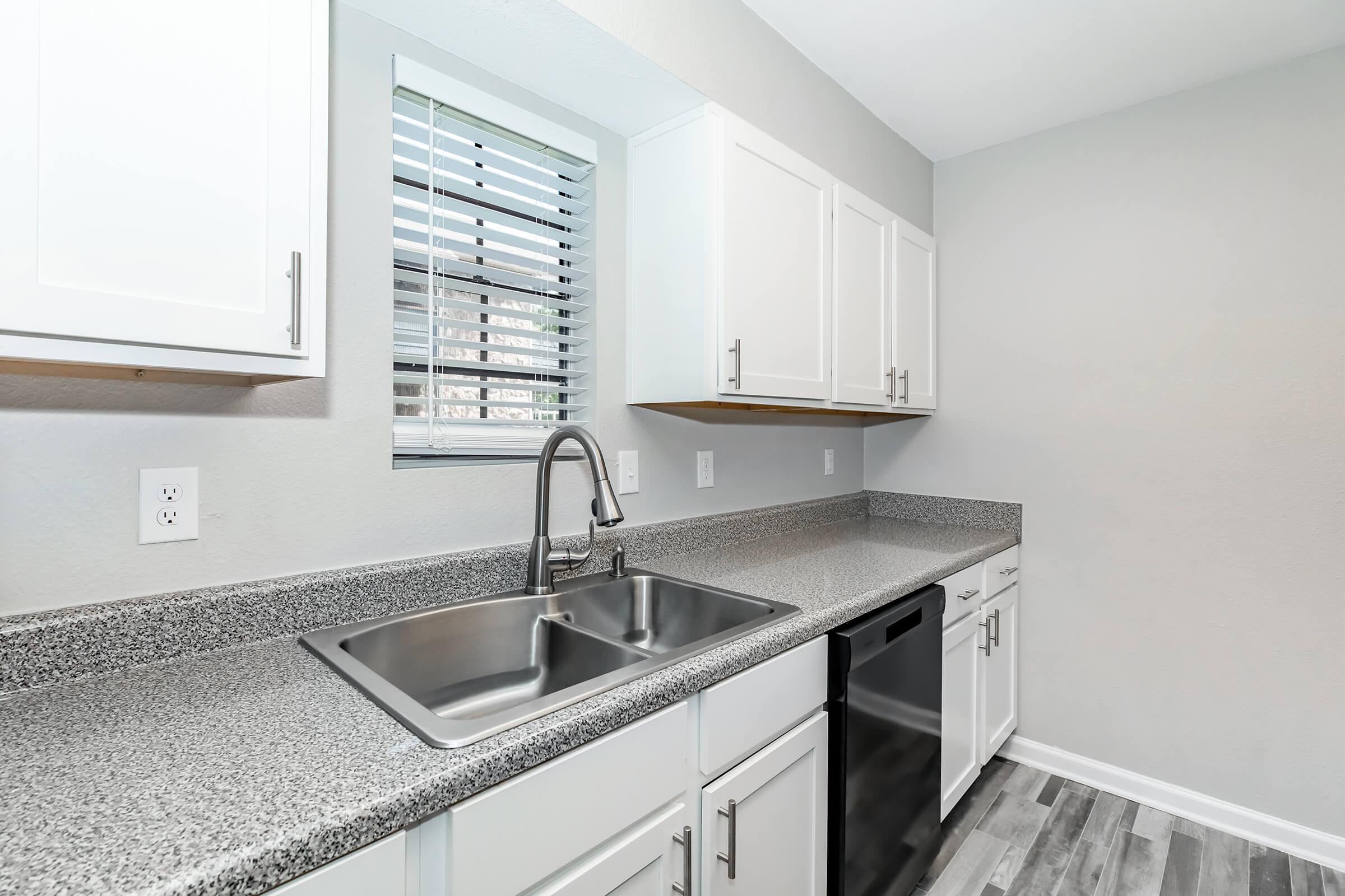 Modern kitchen featuring a gray granite countertop, double sink with a sleek faucet, and white cabinets. A window with blinds provides natural light, and there's a black dishwasher under the counter. The flooring is a light wood-style laminate.