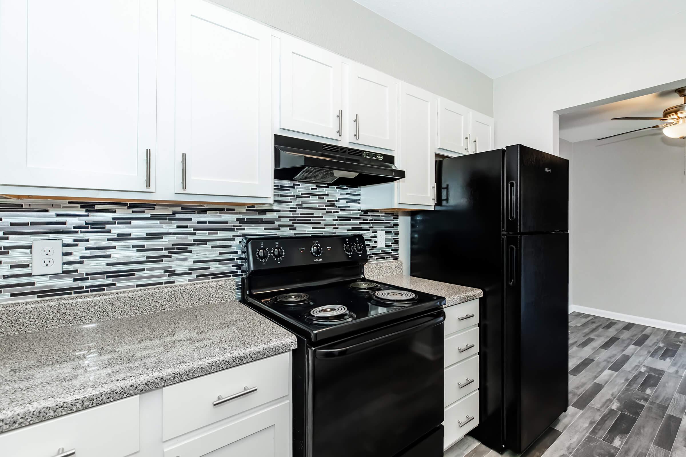 A modern kitchen featuring white cabinetry, a black stove, and a black refrigerator. The backsplash is made up of grey and white tiles, and the countertops are grey with a speckled finish. The floor has a wood-like appearance, and a ceiling fan is visible in the adjacent room.
