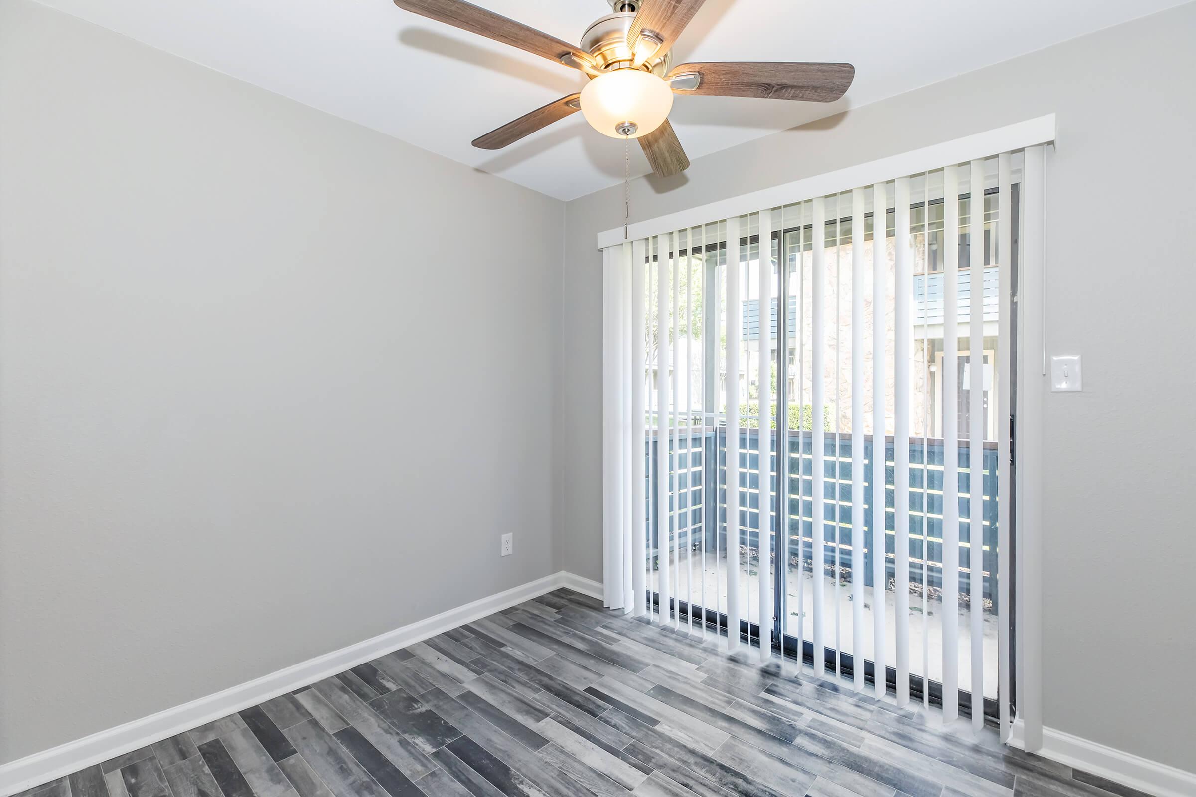 A bright room featuring a ceiling fan, light-colored walls, and modern wood-like flooring. Large sliding glass doors with vertical blinds lead to a small balcony area. The space is well-lit and has a clean, contemporary design, suitable for a living or dining area.