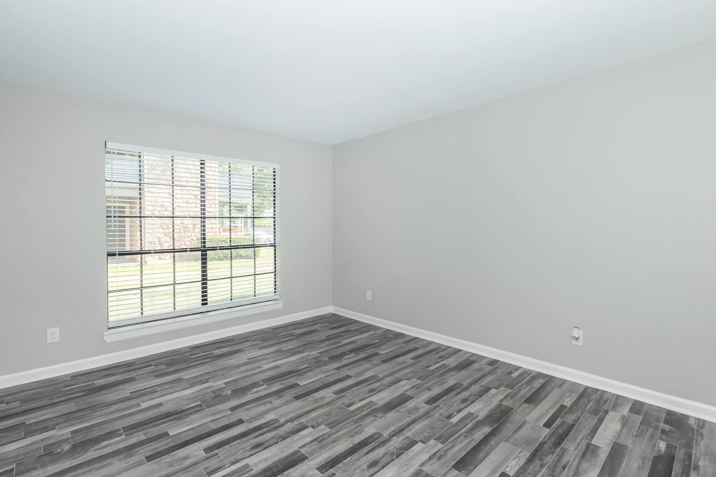 Empty room with light gray walls and gray patterned flooring. A window with white blinds allows natural light to enter. The space appears clean and unfurnished, providing a blank canvas for decoration.