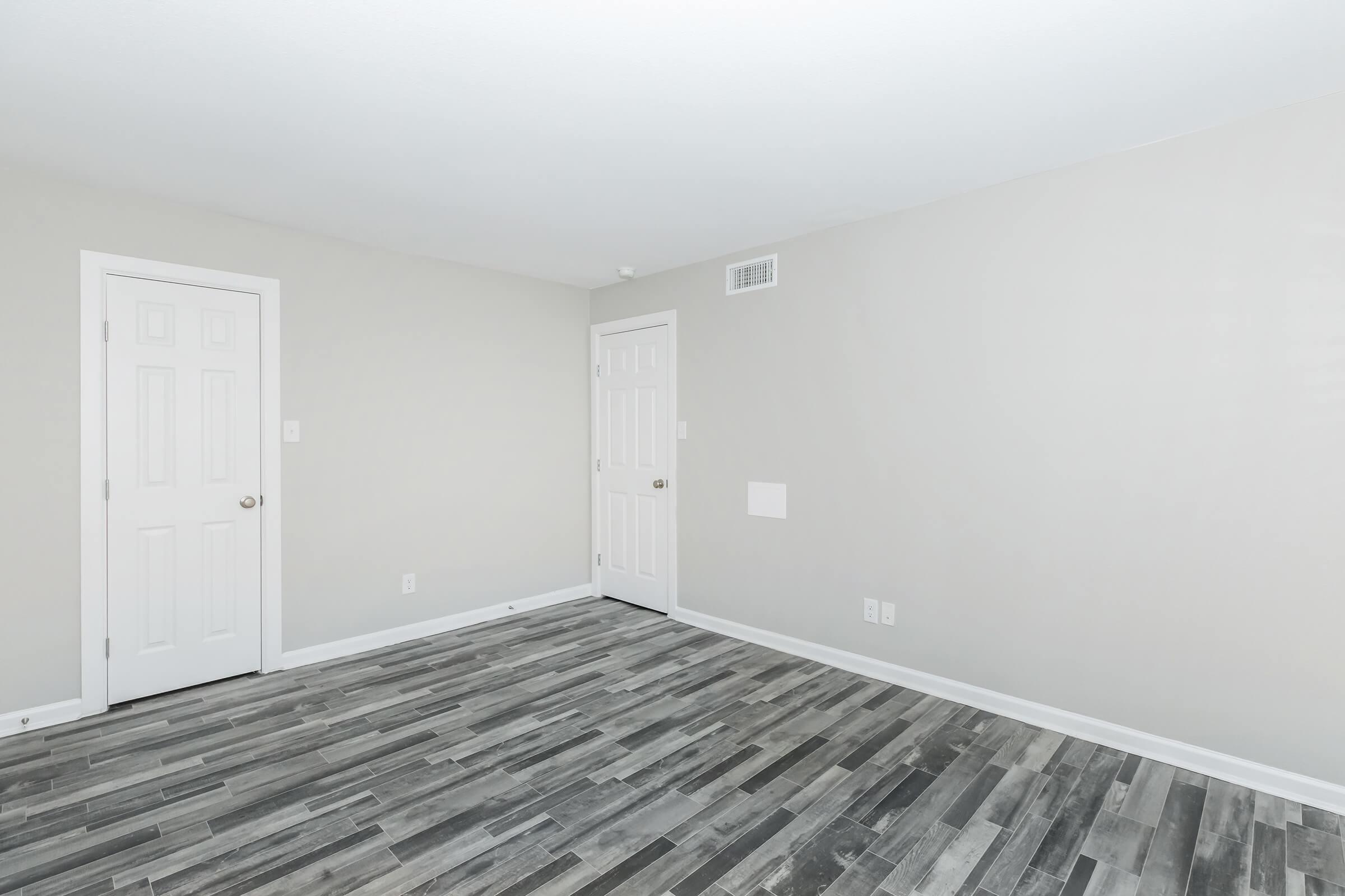 A vacant room featuring light gray walls and a modern wooden floor. Two closed white doors are visible on the left side, and a small wall panel is seen on the right. The room is well-lit, creating a clean and empty appearance, ready for decoration or furnishing.