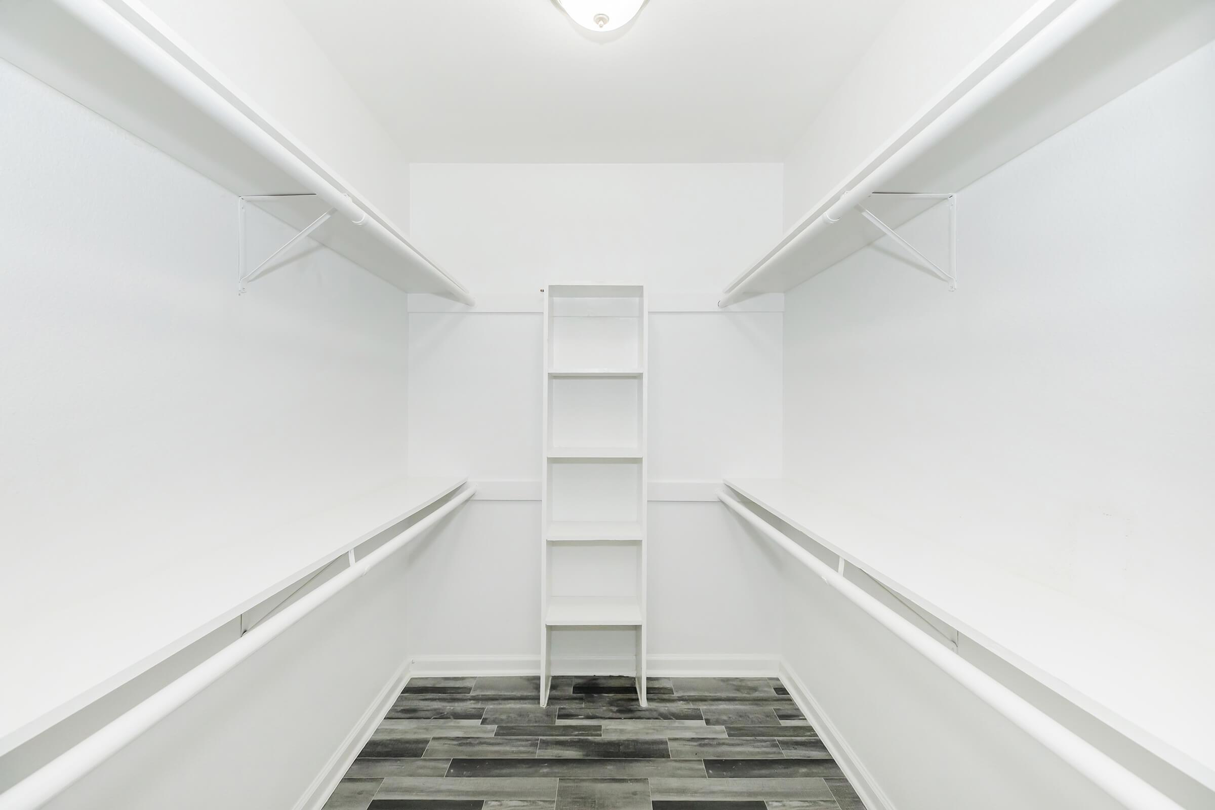 A spacious, empty walk-in closet featuring white walls and shelving. The closet has a central white shelving unit and two long, open hanging areas on either side, with a gray tiled floor. The ceiling is brightened by a light fixture.