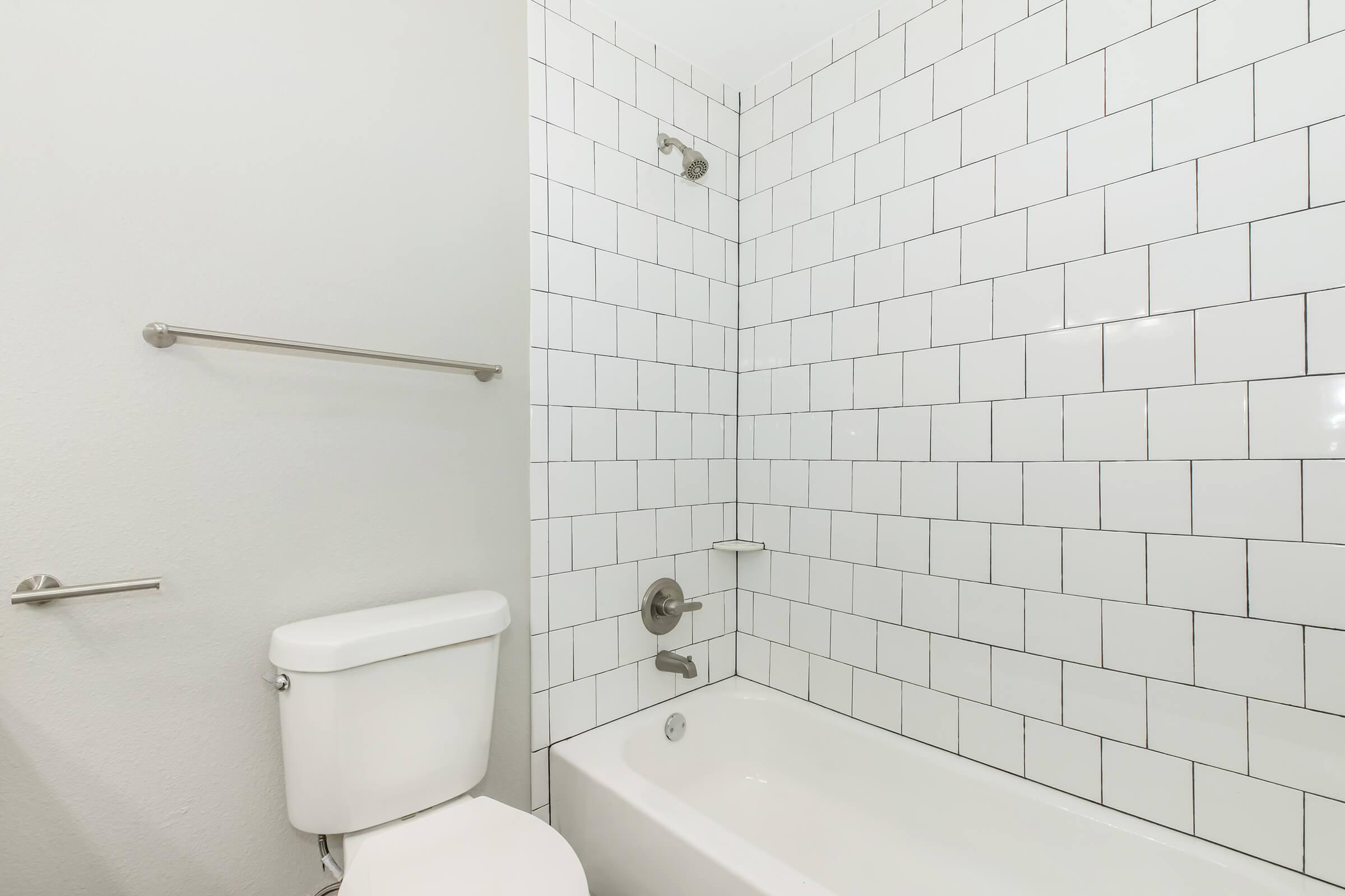 A modern bathroom featuring a white tiled shower area with a bathtub, a toilet, and a chrome towel bar. The walls are painted a light color, creating a clean and spacious feel. The shower fixtures are mounted on the wall, and there's a small shelf built into the tile.