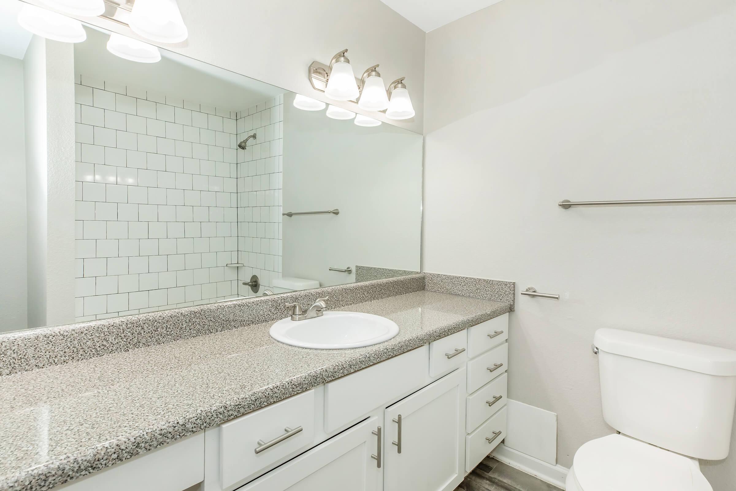 A modern bathroom featuring a white countertop with a sink, a large mirror, and a row of light fixtures above. The walls are painted light gray, and there's a tiled shower area visible in the background. A toilet is positioned to the side, with a clean, minimalistic design.