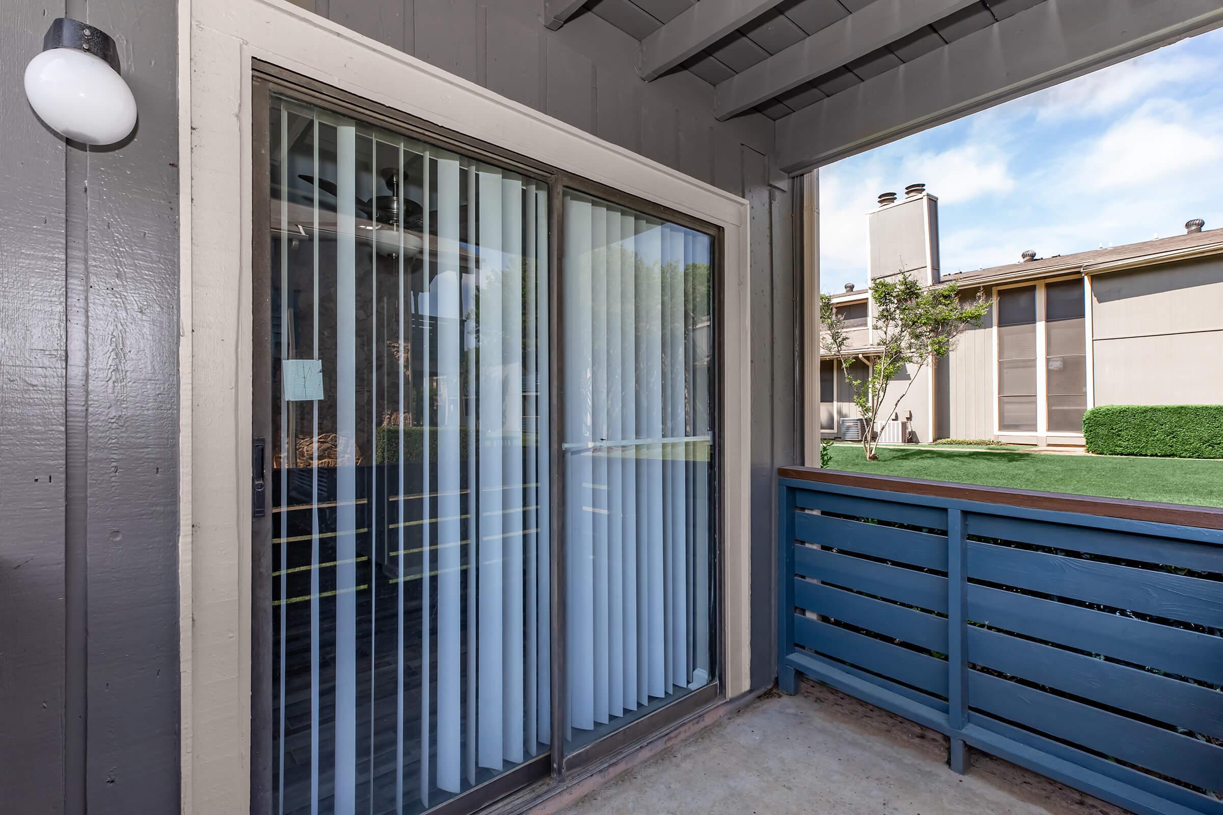 A view of a patio or balcony area featuring sliding glass doors with vertical blinds and a wooden railing. The background shows a green lawn and a building facade. The setting is sunny with a clear blue sky, suggesting a residential environment.