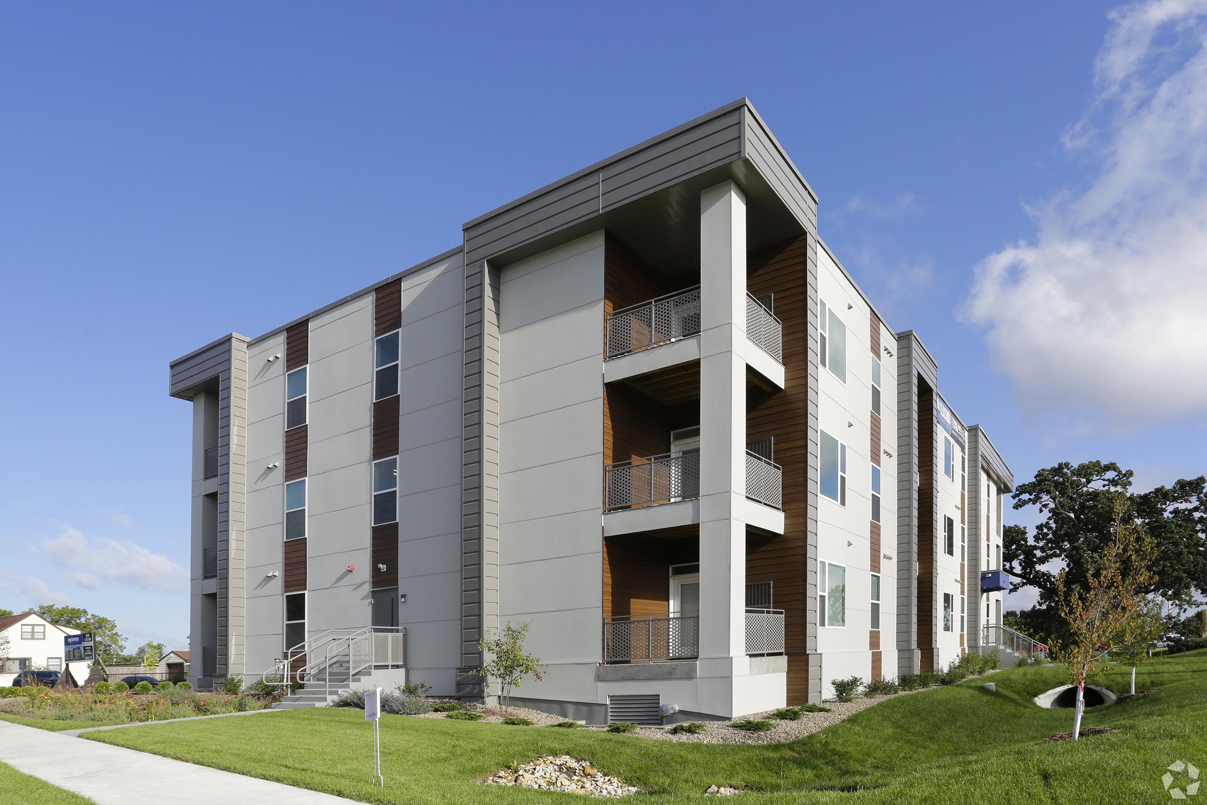 Modern multi-story apartment building with a mix of gray and wooden exterior. Balconies on each floor and landscaped green space in the foreground. Clear blue sky and scattered clouds enhance the bright, inviting atmosphere of the setting.