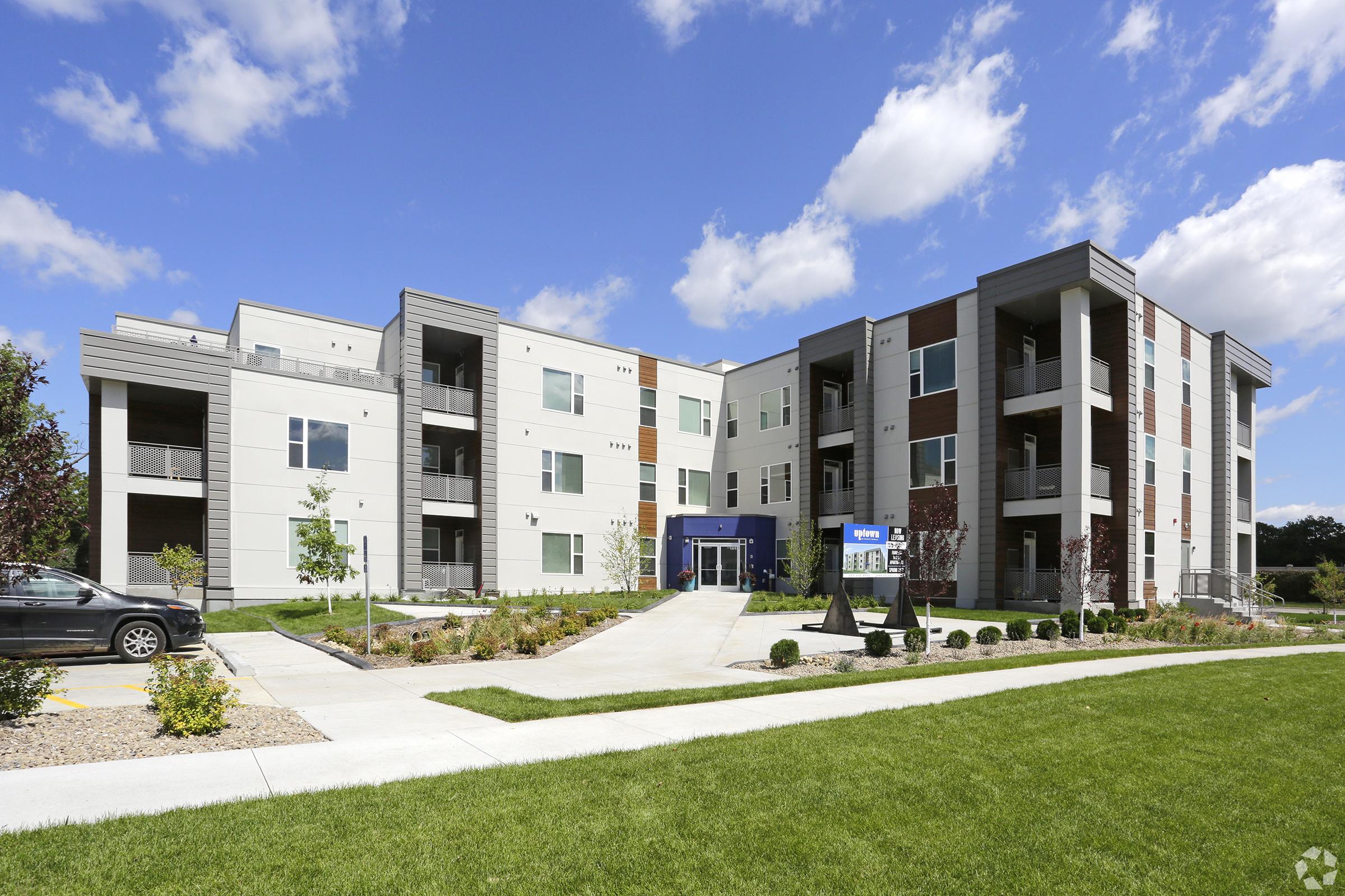 Modern apartment building with a landscaped entrance. Features multiple stories, large windows, and a mix of materials on the facade. The path leads to the main entrance, surrounded by grass and planted shrubs, under a clear blue sky with fluffy clouds.