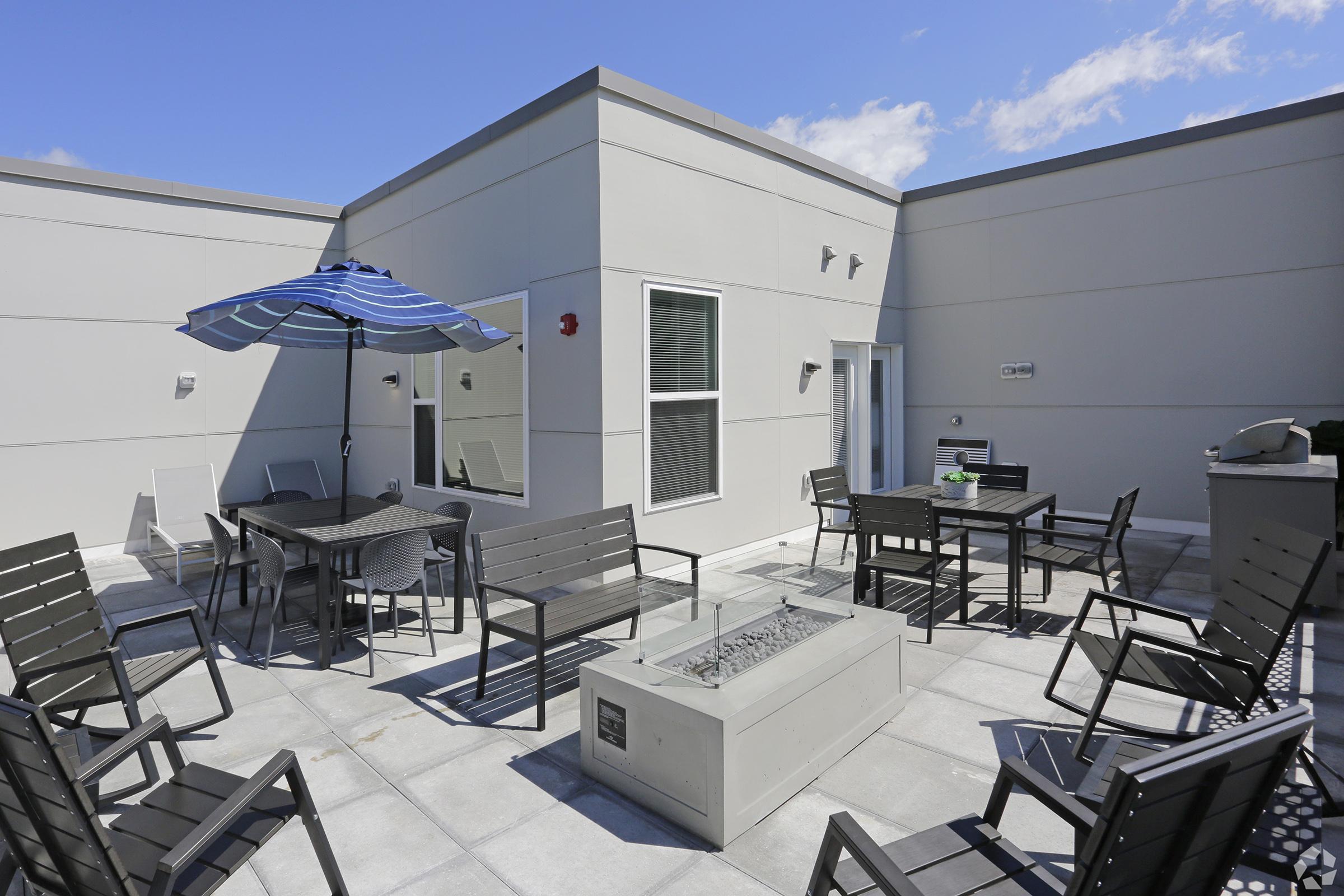 Rooftop terrace featuring modern outdoor furniture, including several black chairs and a table. There's a blue and white striped umbrella for shade, a fire pit in the center, and large, gray walls surrounded by windows. The sky is bright and partly cloudy.