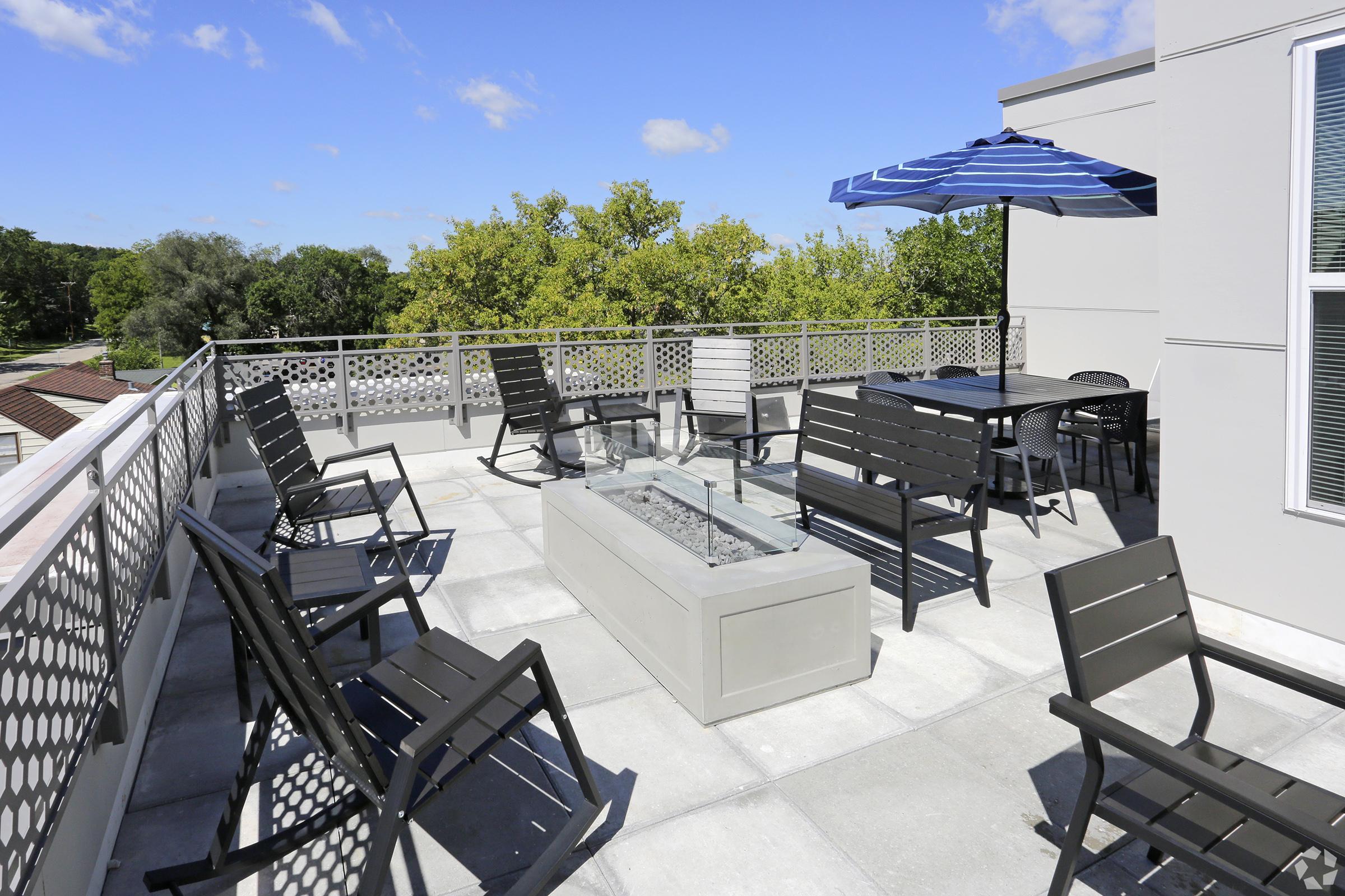 A rooftop patio featuring several black metal chairs and tables, a large blue and white striped umbrella, and a central fire table filled with decorative stones. Lush greenery is visible in the background under a clear blue sky.