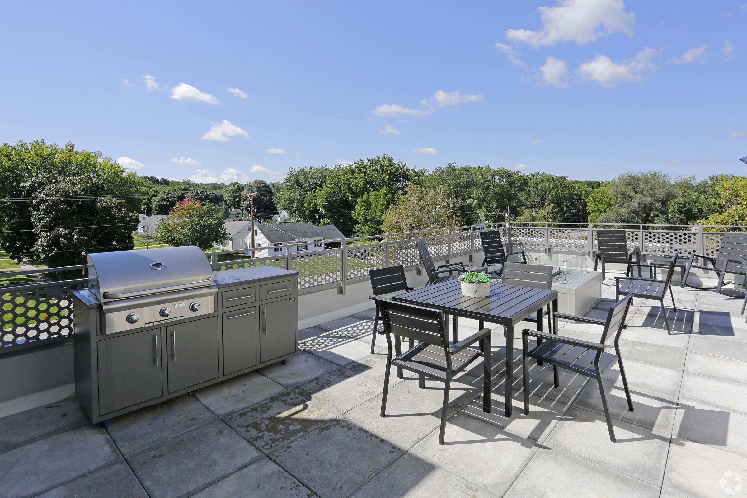 A spacious rooftop terrace featuring a modern grill, a dining table with several chairs, and a panoramic view of green trees and houses under a clear blue sky. Sunlight illuminates the area, creating a welcoming outdoor space for gatherings and relaxation.