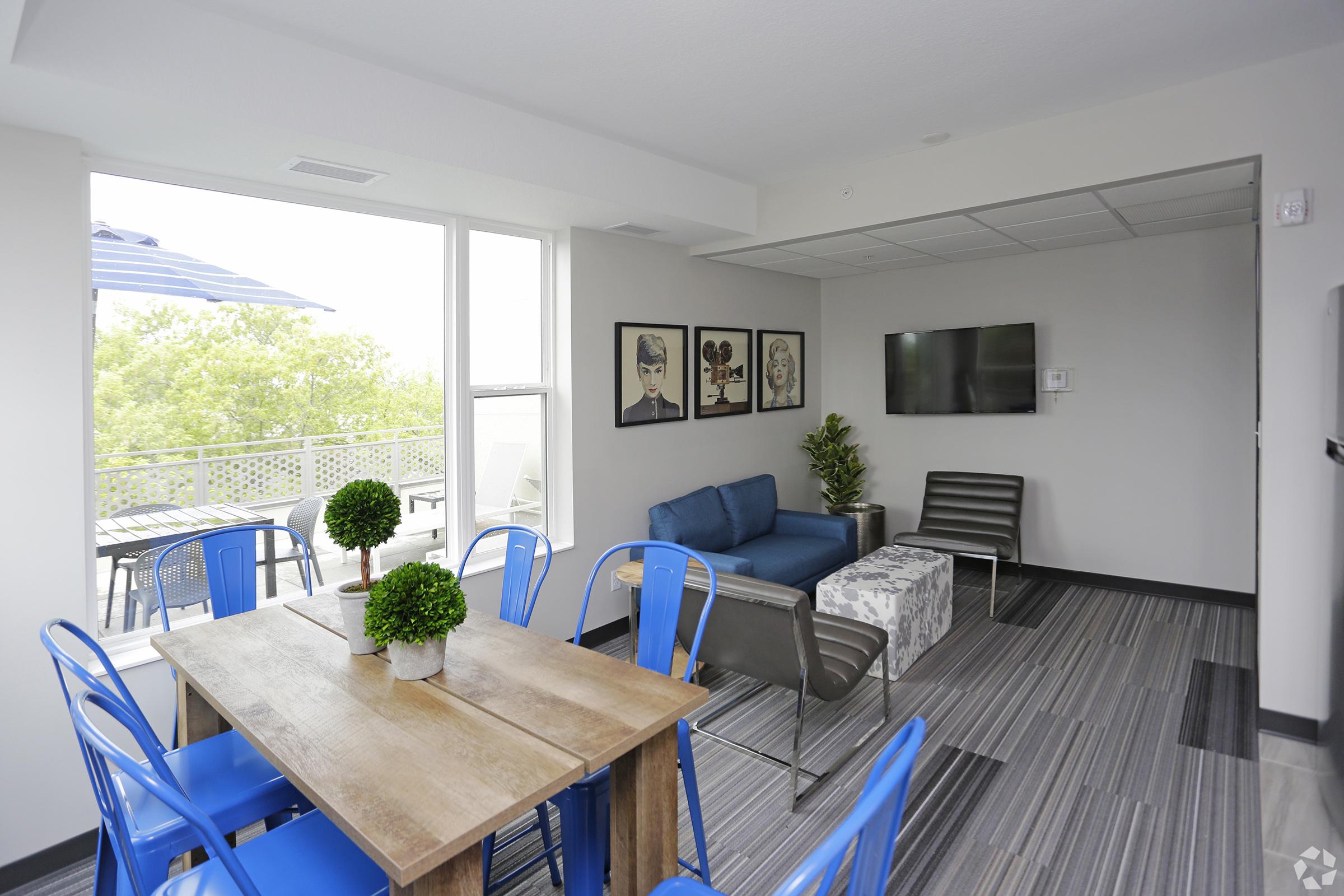 A modern lounge area featuring a blue couch, two chairs, and a wooden dining table with blue chairs. Three framed artworks hang on the wall. A flat-screen TV is mounted opposite the seating. Large windows provide natural light, overlooking a patio with an umbrella.