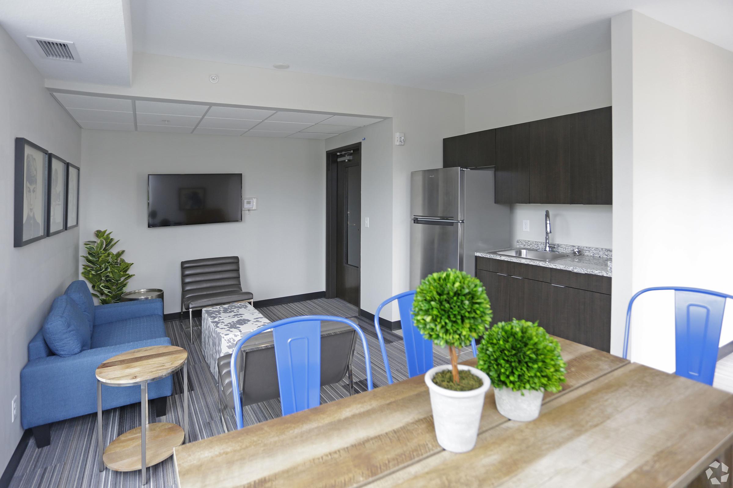 Interior of a modern apartment featuring a living area with a blue sofa, a gray armchair, and a small coffee table. A dining table with blue chairs is in the foreground. The kitchen area includes stainless steel appliances and dark wood cabinets. Decorative plants add a touch of greenery to the space.