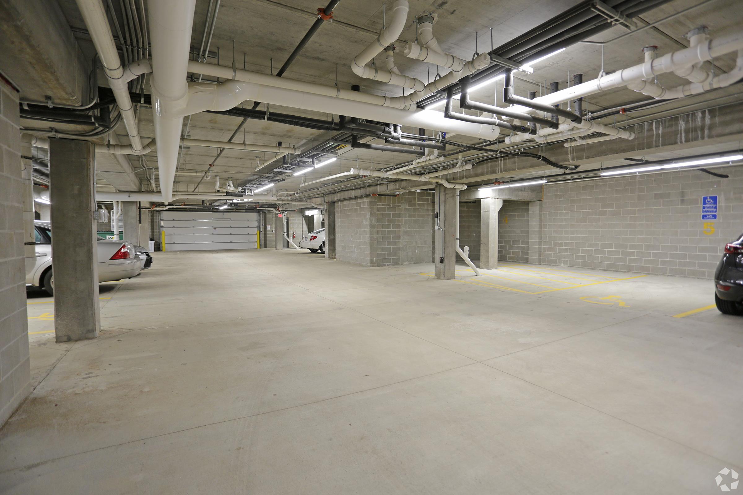 A spacious underground parking garage with concrete floors, gray block walls, and overhead lighting. Several parking spaces are marked, and a few cars are visible parked along the sides. The area appears clean and well-maintained, with pipes and electrical fixtures running along the ceiling.