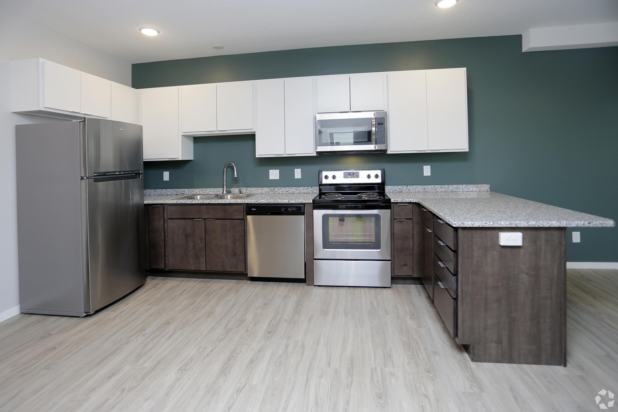 Modern kitchen featuring stainless steel appliances, including a refrigerator, microwave, and stove. The cabinets are a mix of white and dark wood, with a sleek granite countertop. The walls are painted teal, and the flooring is light-colored laminate, creating a bright and inviting atmosphere.