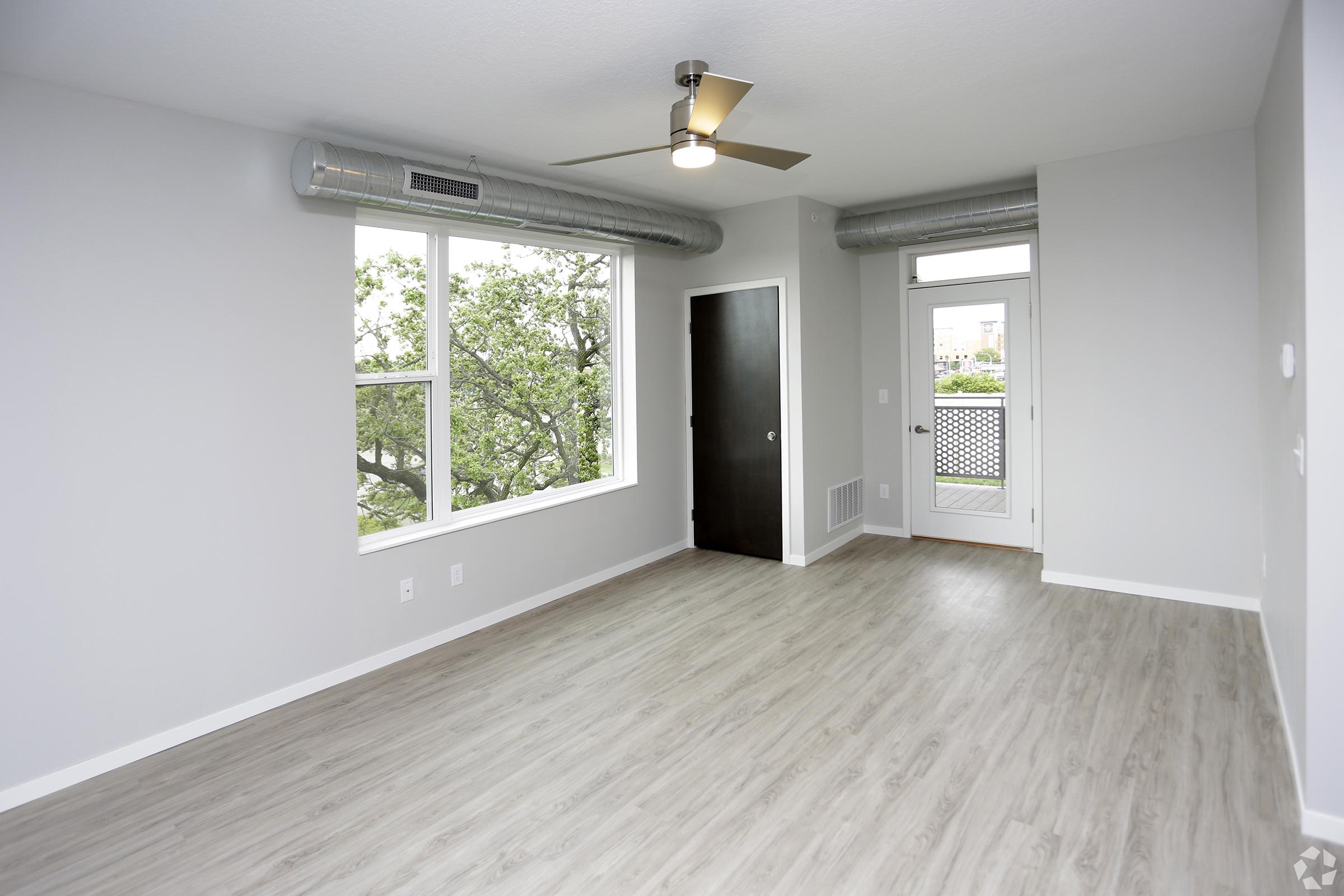 Empty room with light gray walls and wooden flooring. Large windows allow natural light to enter, showcasing greenery outside. A ceiling fan is installed, and there is a door leading to a balcony. The room has an open layout, emphasizing spaciousness. No furniture or decor is present.