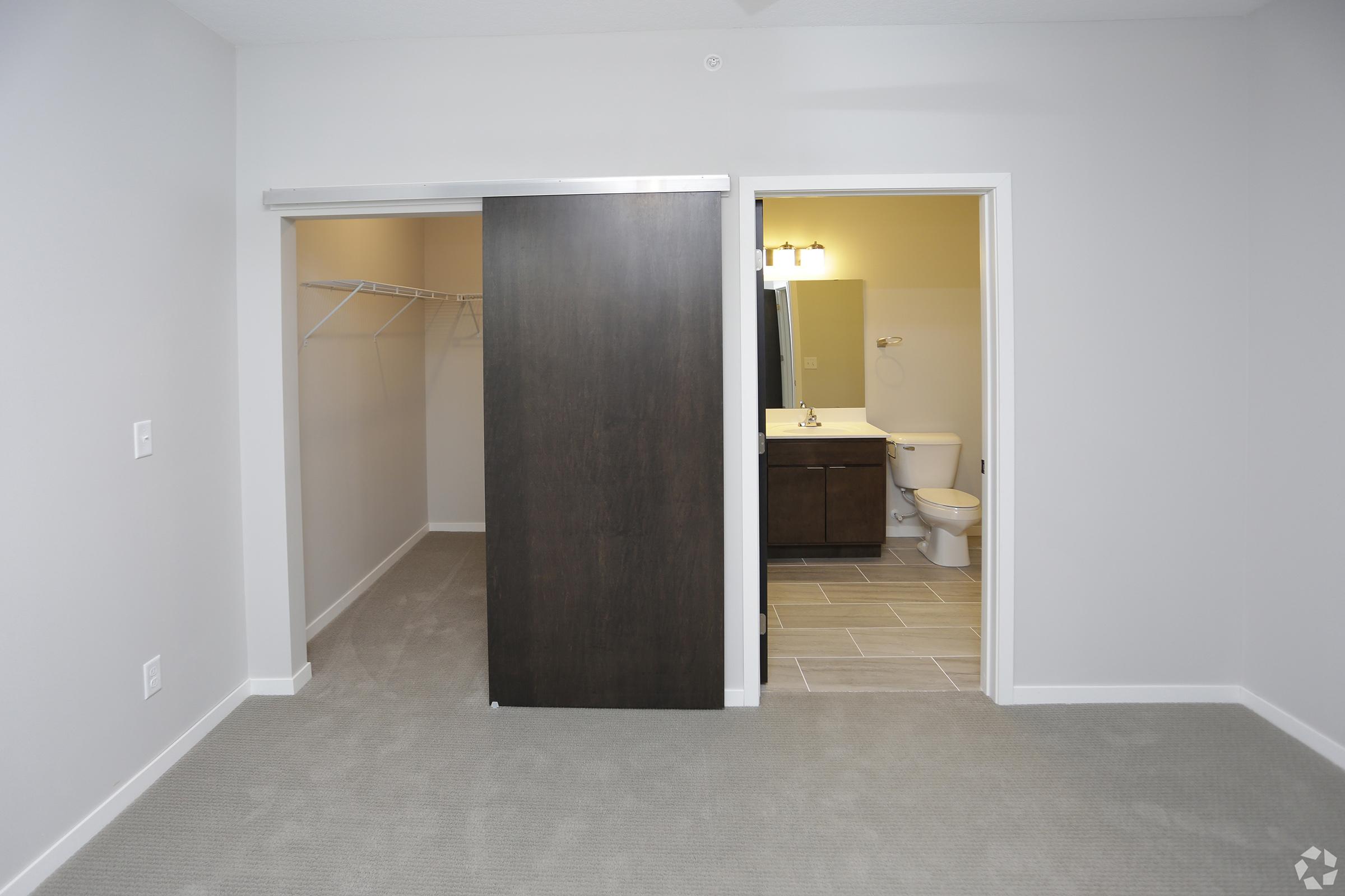 A spacious room featuring a sliding closet door on the left, revealing a simple, empty closet. To the right, a doorway leads to a bathroom with a dark vanity and a toilet, complemented by light-colored tile flooring. The walls are painted in neutral tones, enhancing the open and airy feel of the space.