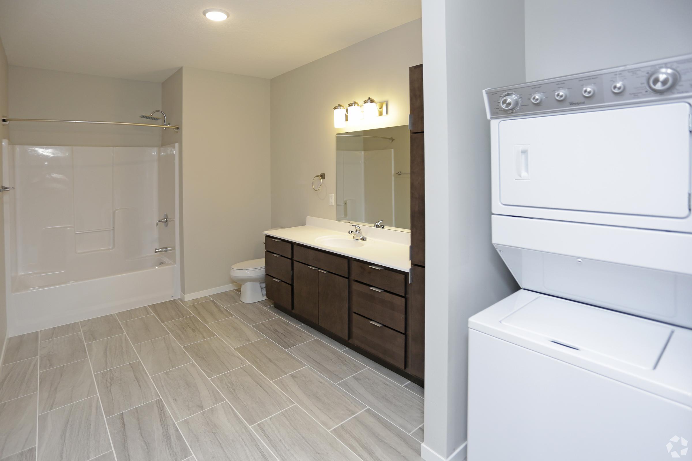A modern bathroom featuring a white bathtub with a shower, a large mirror above a dark wood double vanity with sinks, and a washer and dryer unit in a corner. The space has gray tiled flooring and neutral-colored walls, creating a clean and contemporary atmosphere.