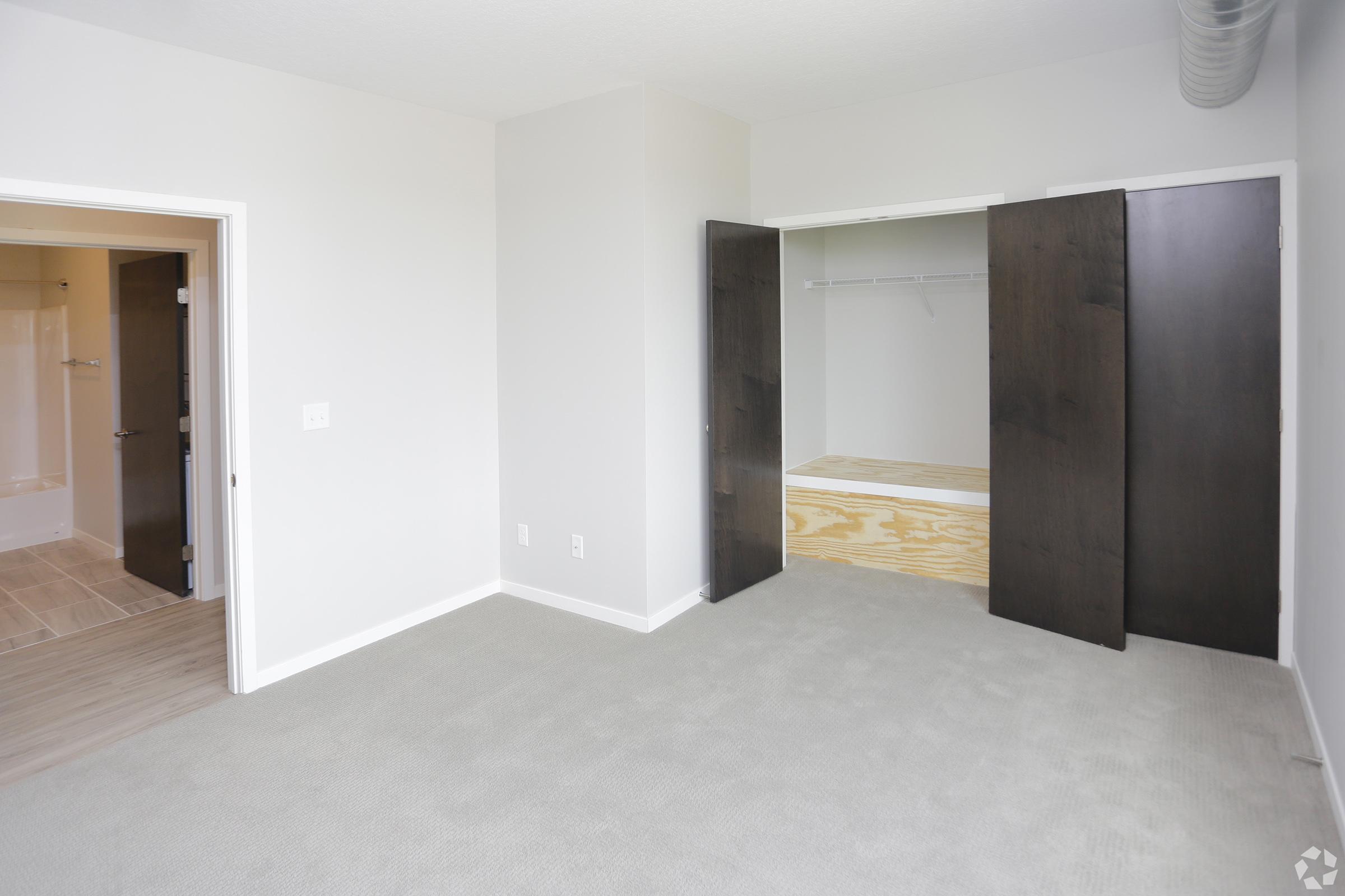 A spacious, light-filled room with beige carpet flooring and light gray walls. There is an open closet with wooden shelving and two dark wooden doors leading to it. A doorway in the background leads to a small bathroom area, but only a glimpse is visible. The overall atmosphere is clean and modern.