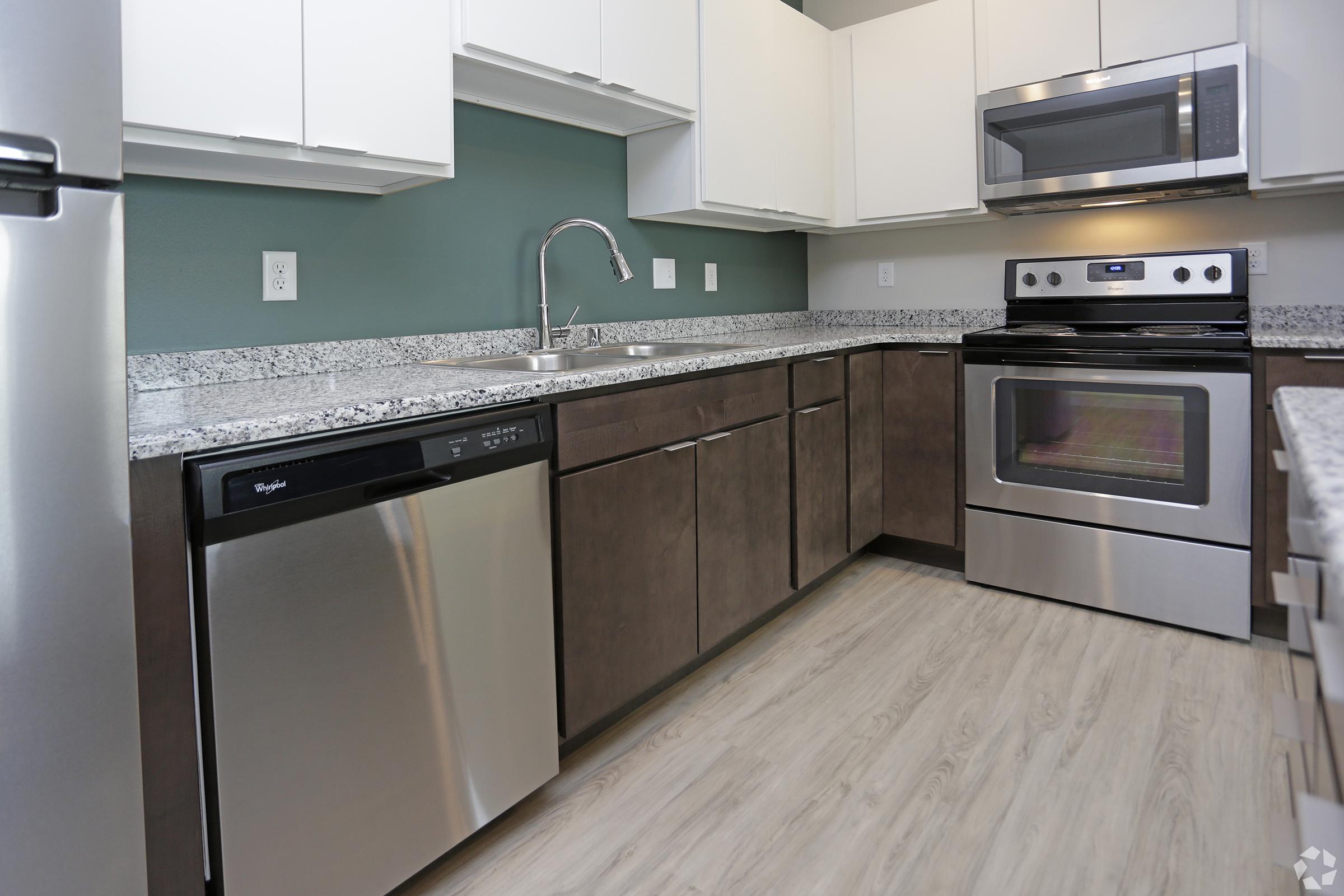 Modern kitchen featuring stainless steel appliances including a dishwasher, microwave, and oven. The cabinetry is a mix of light and dark wood, with a light granite countertop and a green accent wall. The kitchen is organized and well-lit, showcasing a clean, contemporary design.