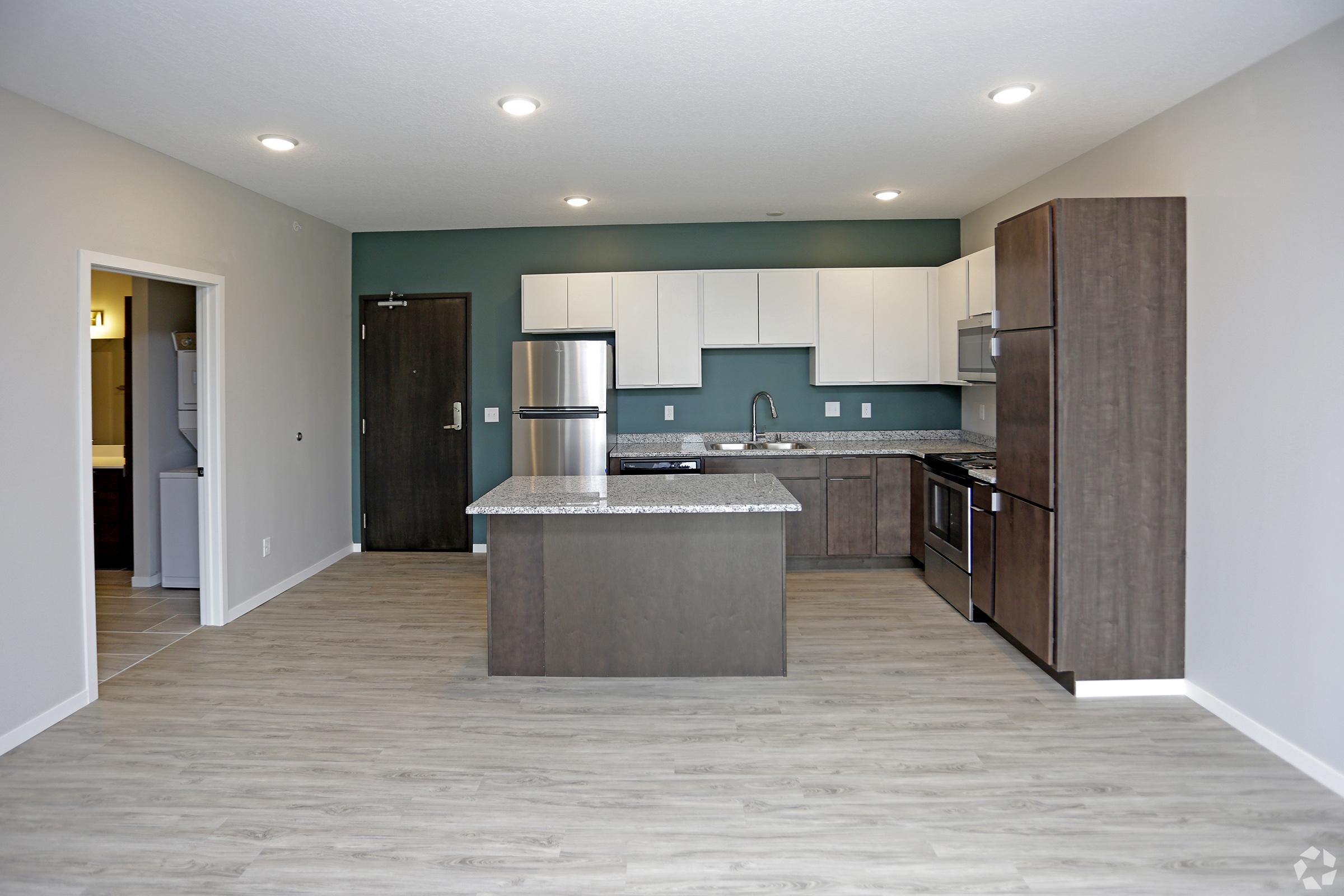 A modern kitchen featuring light wood flooring, an island with a granite countertop, stainless steel appliances, and white cabinetry. The space is well-lit with recessed lighting, and a dark green accent wall adds contrast. An open layout connects the kitchen to the living area, displaying a contemporary design.
