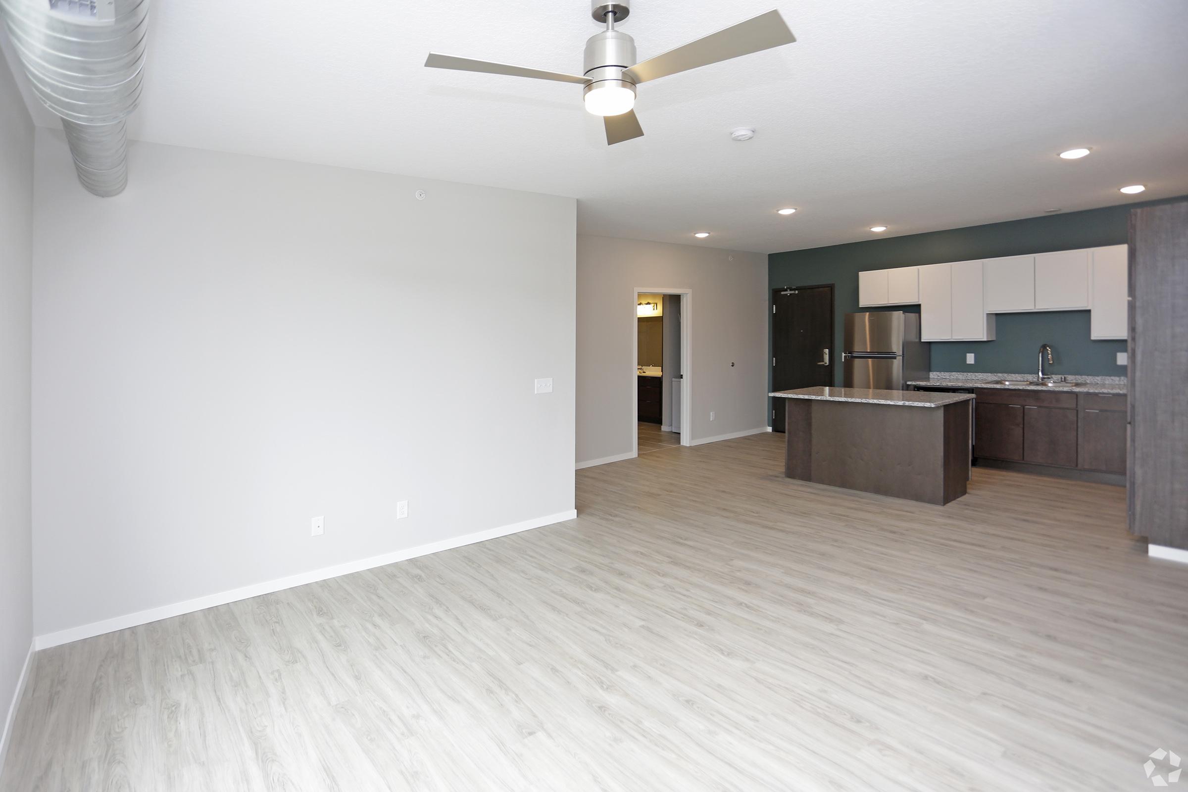 Spacious modern living area with light wood flooring, a ceiling fan, and an open concept layout. The adjacent kitchen features dark cabinets, stainless steel appliances, and a large island. Walls are painted in neutral tones, enhancing the airy feel of the space.