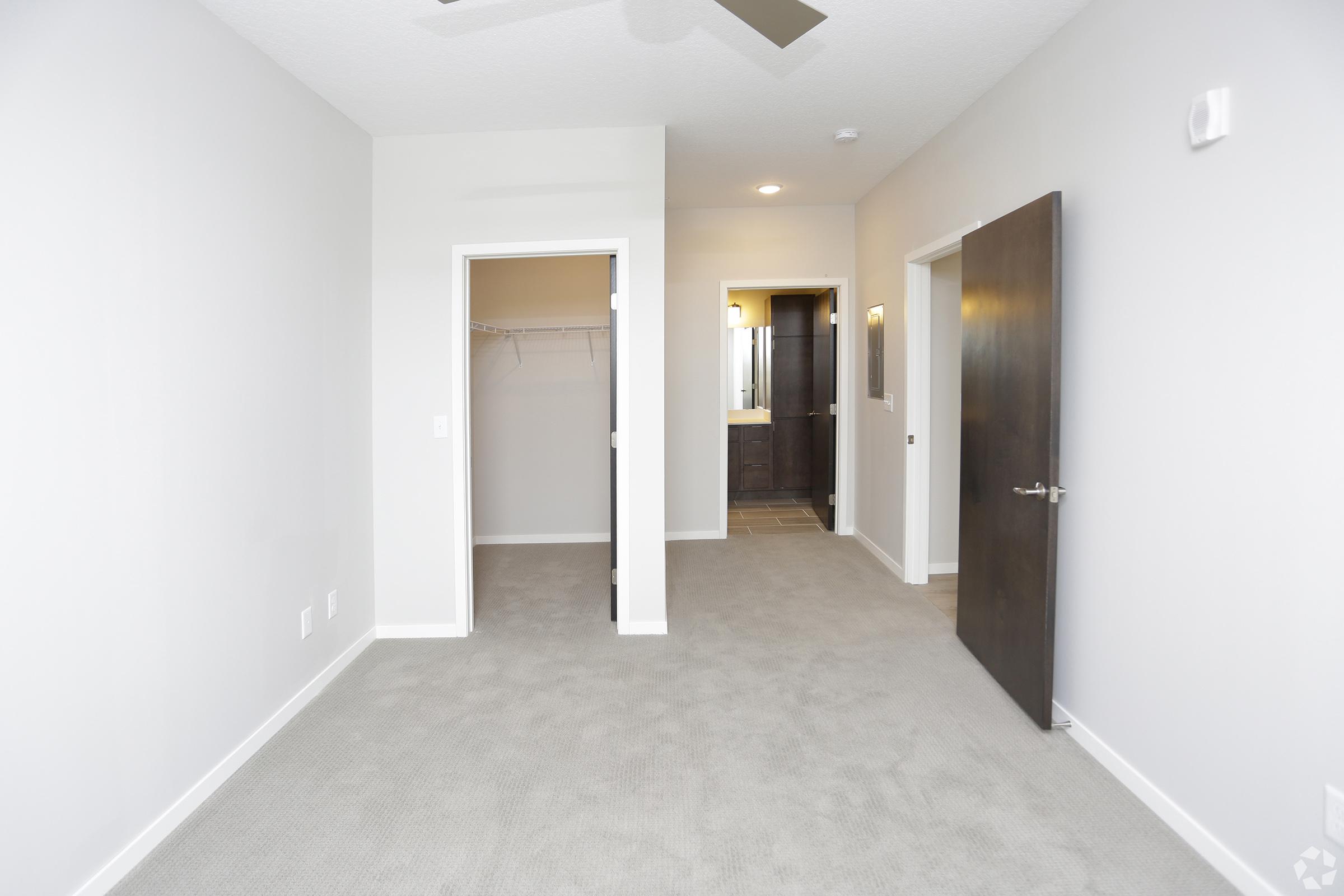 A spacious, empty room with light gray walls and carpeted flooring. Two doorways lead to a closet on the left and a bathroom on the right, which is partially visible. A ceiling fan hangs from the ceiling, and warm light fixtures are mounted on the walls. The overall atmosphere is bright and modern.