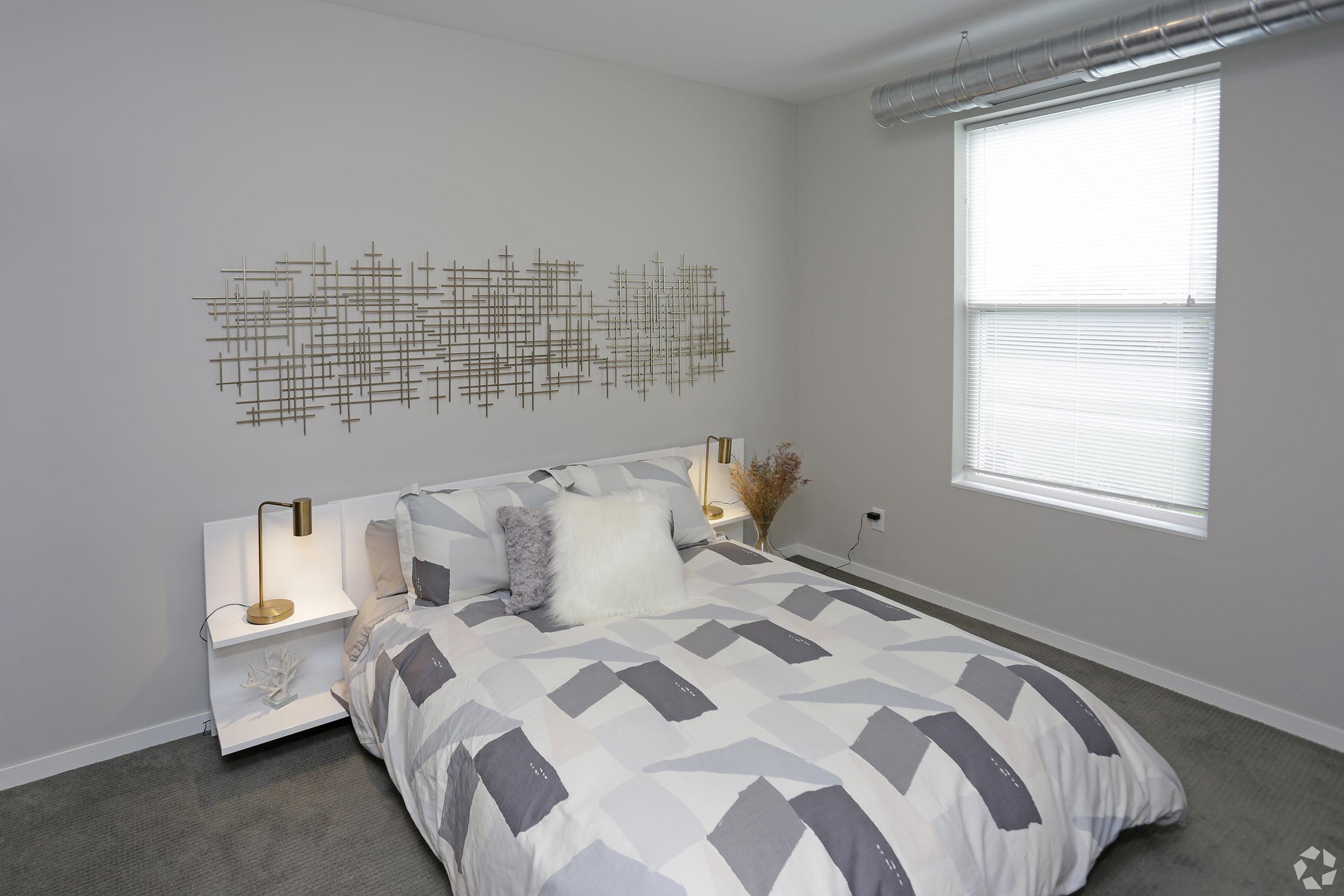 A modern bedroom featuring a bed with a patterned gray and white comforter, fluffy pillows, and two bedside lamps. The walls are light gray, and there is a decorative wall art piece made of gold geometric shapes above the bed. A window provides natural light, and a potted plant adds a touch of greenery.