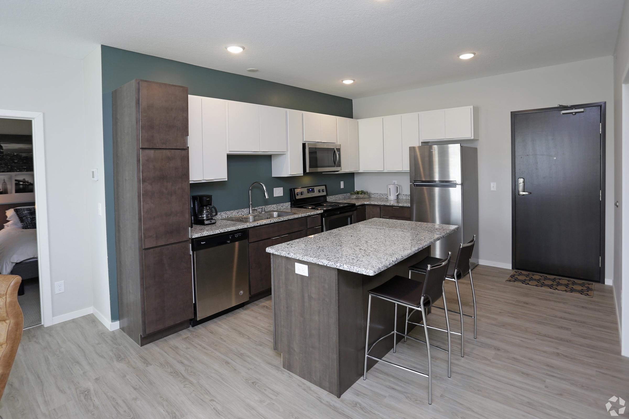 Modern kitchen featuring dark wood cabinets, stainless steel appliances, and a granite countertop island with bar stools. Light-colored flooring and a blue accent wall enhance the contemporary design. The kitchen opens to a living area, with a door leading to an exterior space.