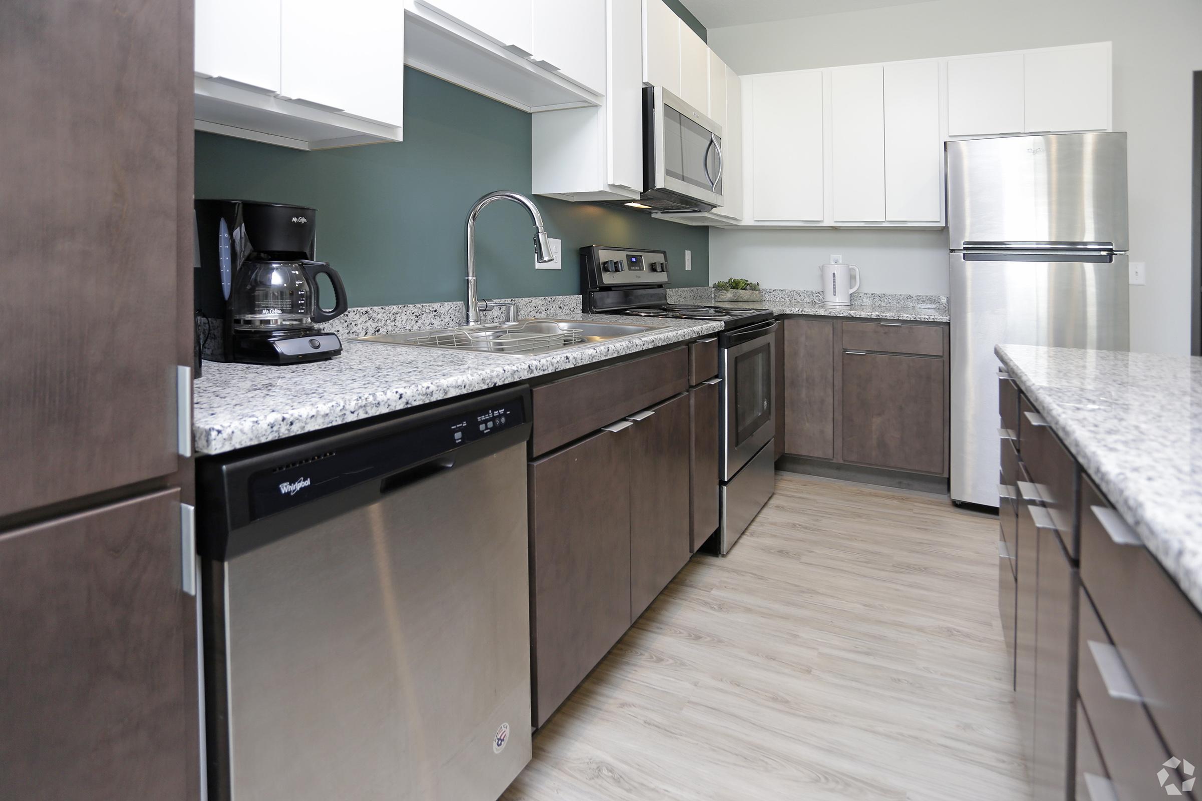 Modern kitchen featuring stainless steel appliances, including a refrigerator, oven, and microwave. There is a black coffee maker on the counter, and the cabinets are a mix of light and dark finishes. The countertop is made of speckled granite, and the floor is light wood. The walls are painted in a soothing green color.
