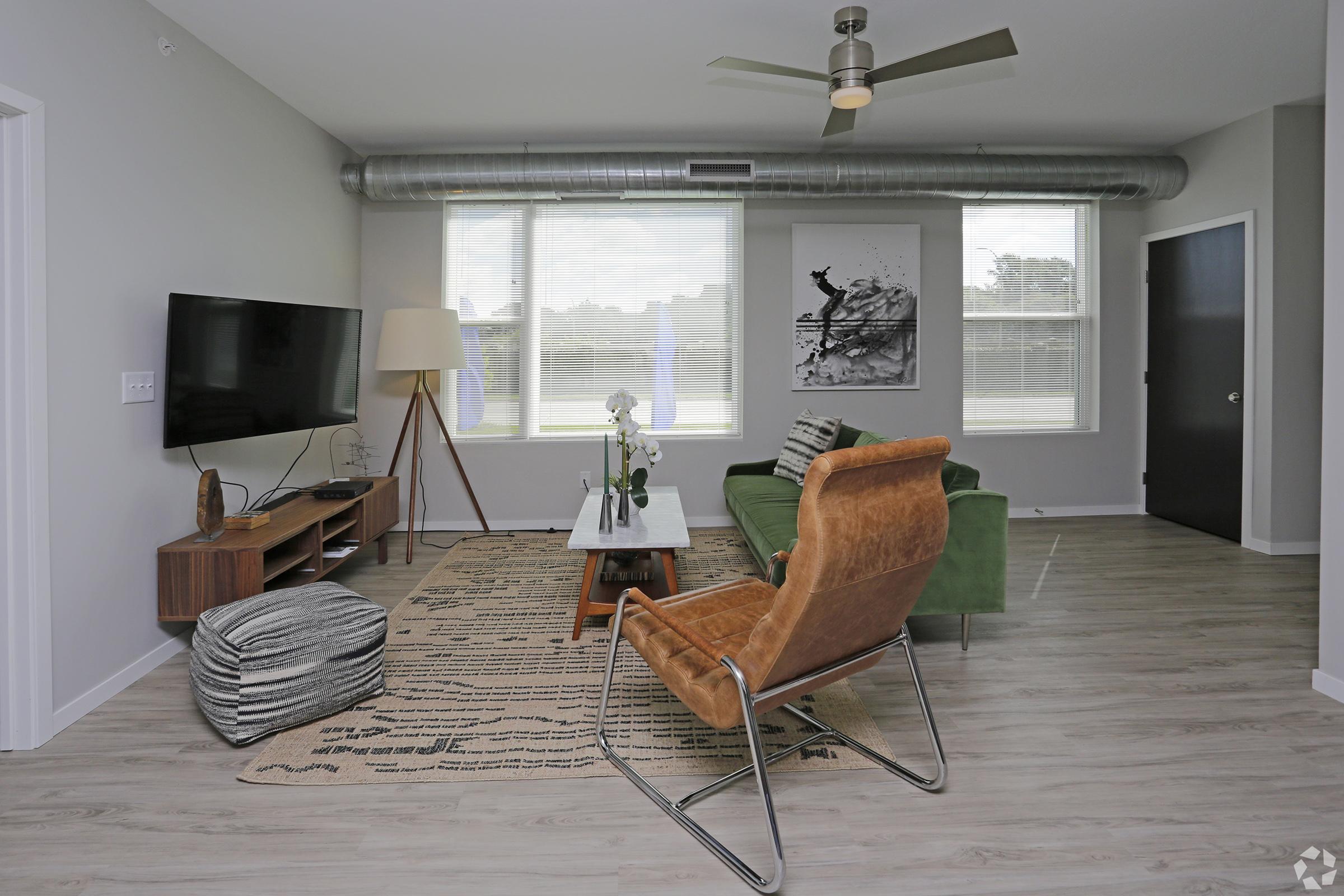 A modern living room featuring a green sofa, a brown leather chair, and a wooden coffee table. There is a television on a wooden stand, a decorative floor lamp, and a large window allowing natural light in. The walls are light-colored, and a piece of artwork is displayed on one wall.