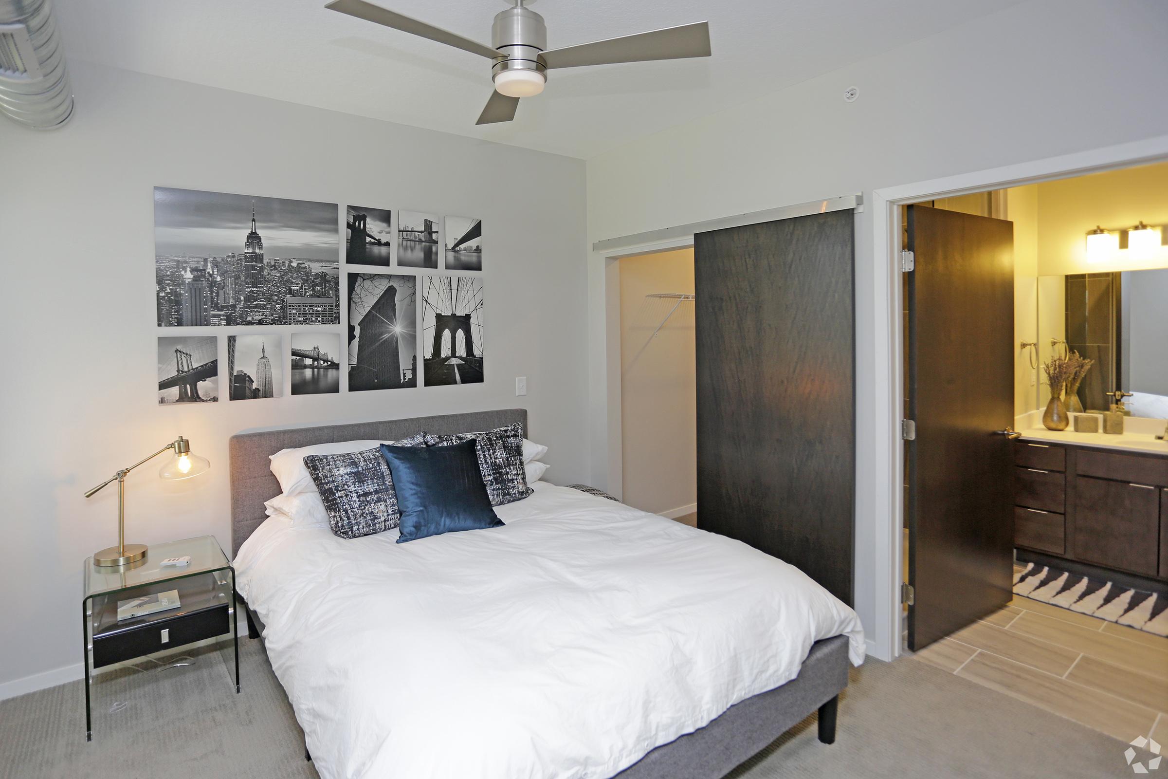 A cozy bedroom featuring a queen-sized bed with white bedding and decorative pillows. The wall displays black-and-white photographs of a city skyline. A modern lamp sits on a nightstand, and a bathroom is visible through an open door with dark wooden accents. A ceiling fan adds a contemporary touch.