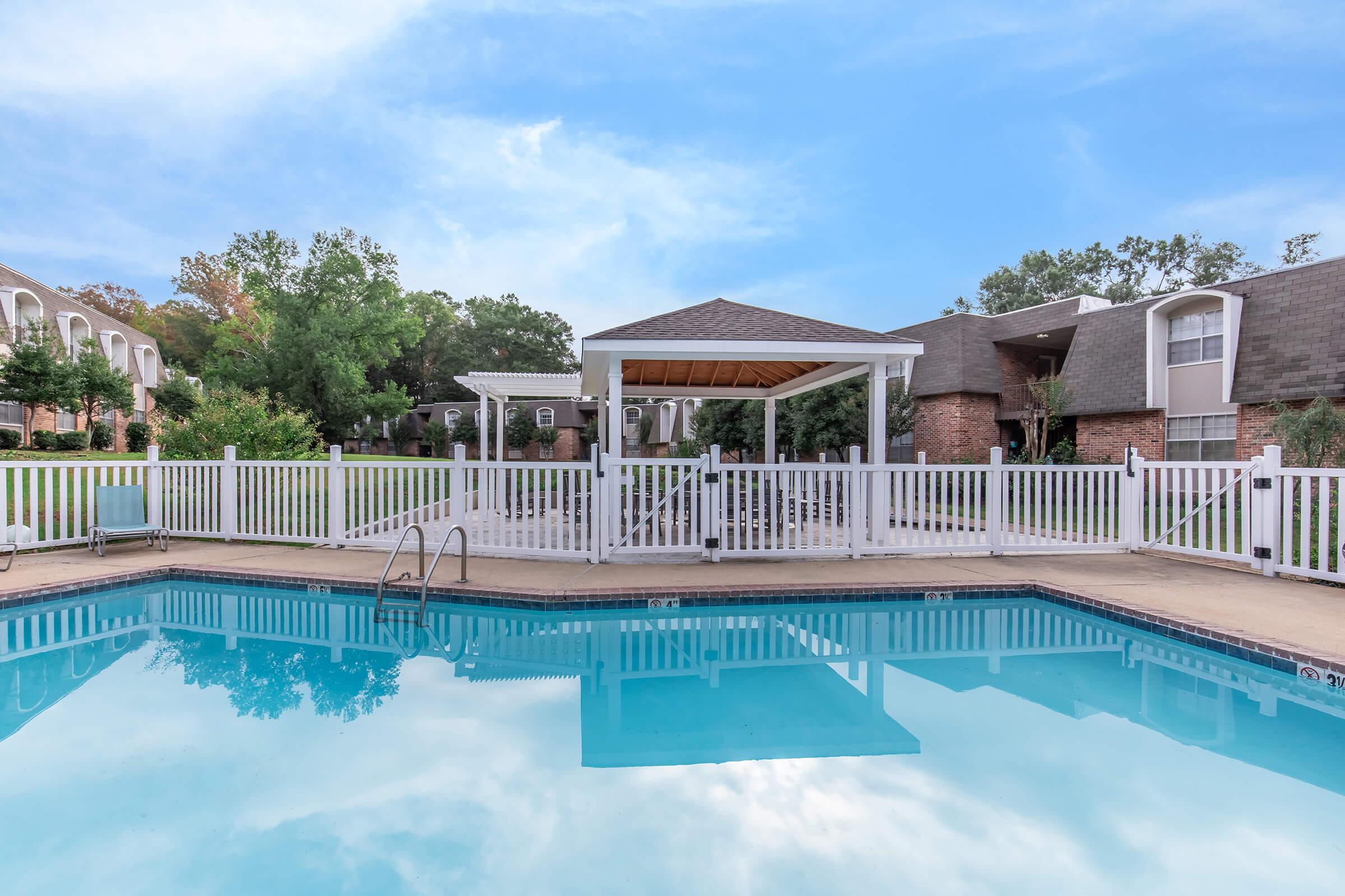 A clear blue swimming pool surrounded by a white fence, with a gazebo in the background. Green trees and landscaped grounds are visible, along with nearby residential buildings under a bright blue sky. The serene atmosphere invites relaxation and leisure activities.