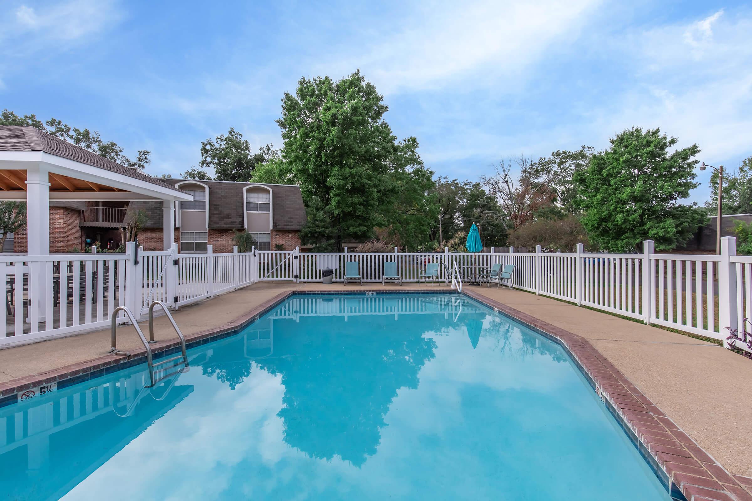 A clear swimming pool surrounded by a white picket fence, with a gazebo in the background. Lush green trees and a bright blue sky enhance the tranquil outdoor setting. The pool deck features a ladder for entry, and there are lounge chairs nearby, inviting relaxation.