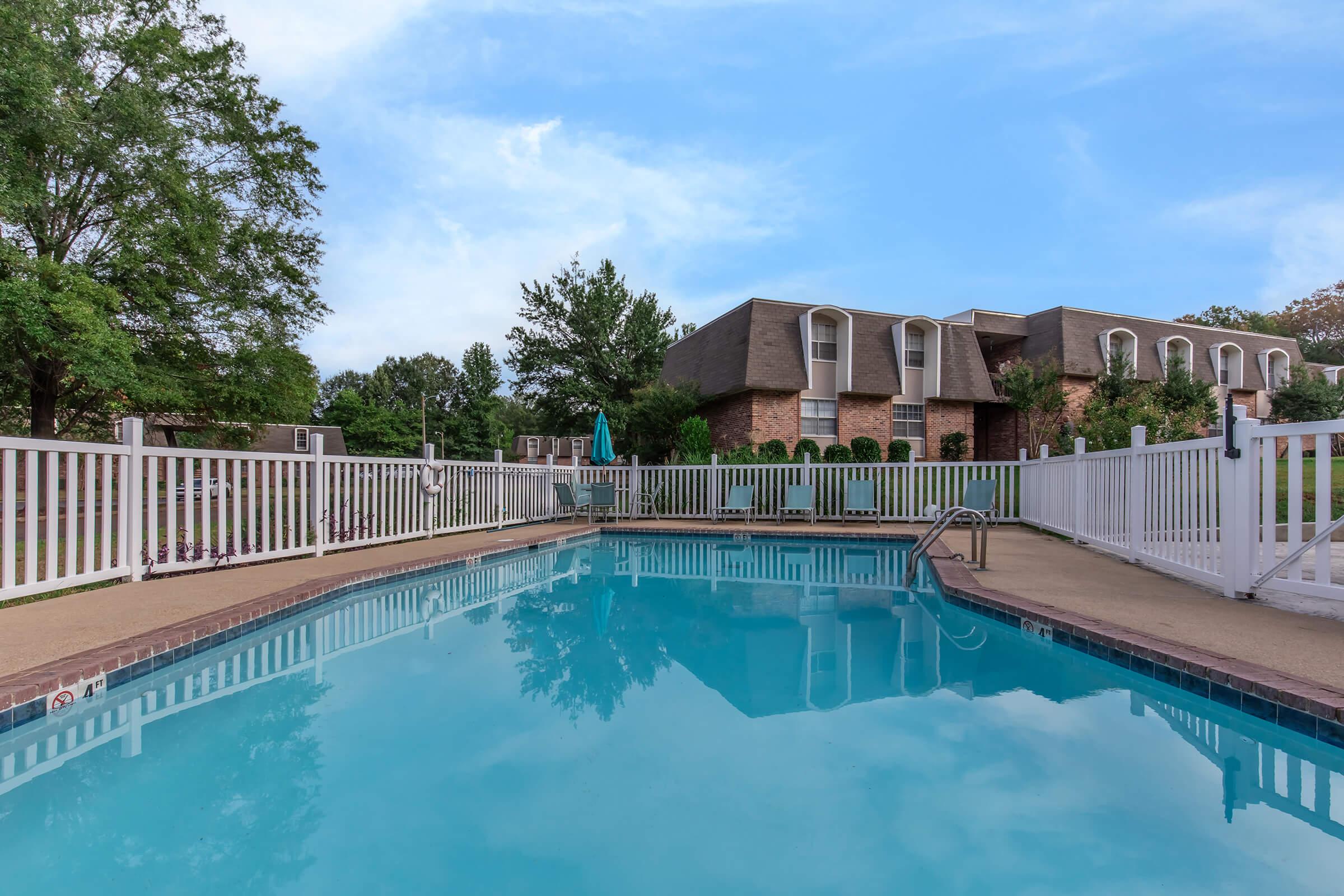 A clear swimming pool surrounded by a white fence, featuring a ladder for entry. In the background, there is a brick building with a triangular roof and several trees. The sky is bright blue with scattered clouds, creating a serene outdoor atmosphere.