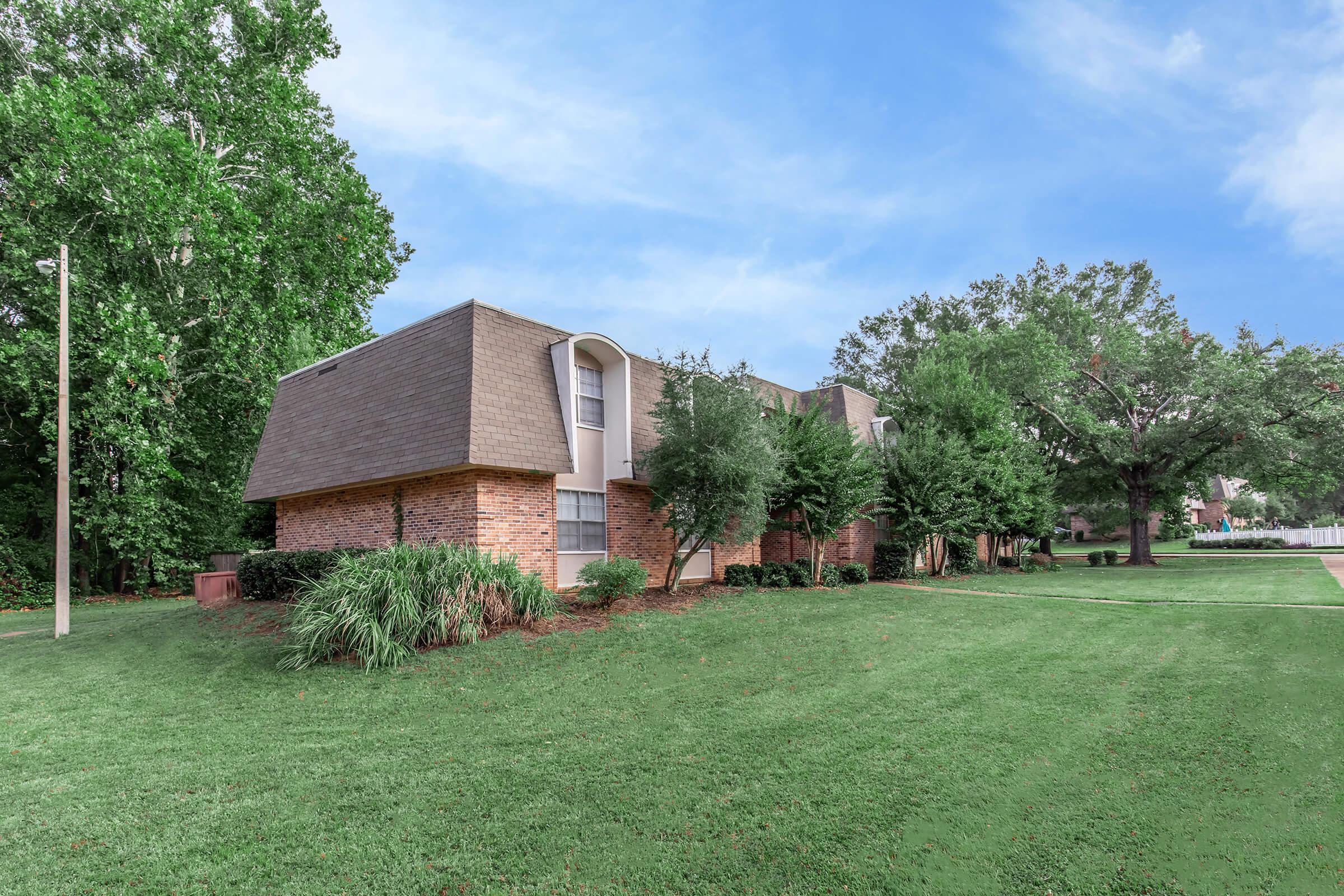 A landscape view of a modern building featuring a sloped roof, made of brick and surrounded by well-maintained greenery. Lush trees and grass are visible in the foreground, with a clear blue sky in the background. The setting is serene and inviting, typical of a residential area.