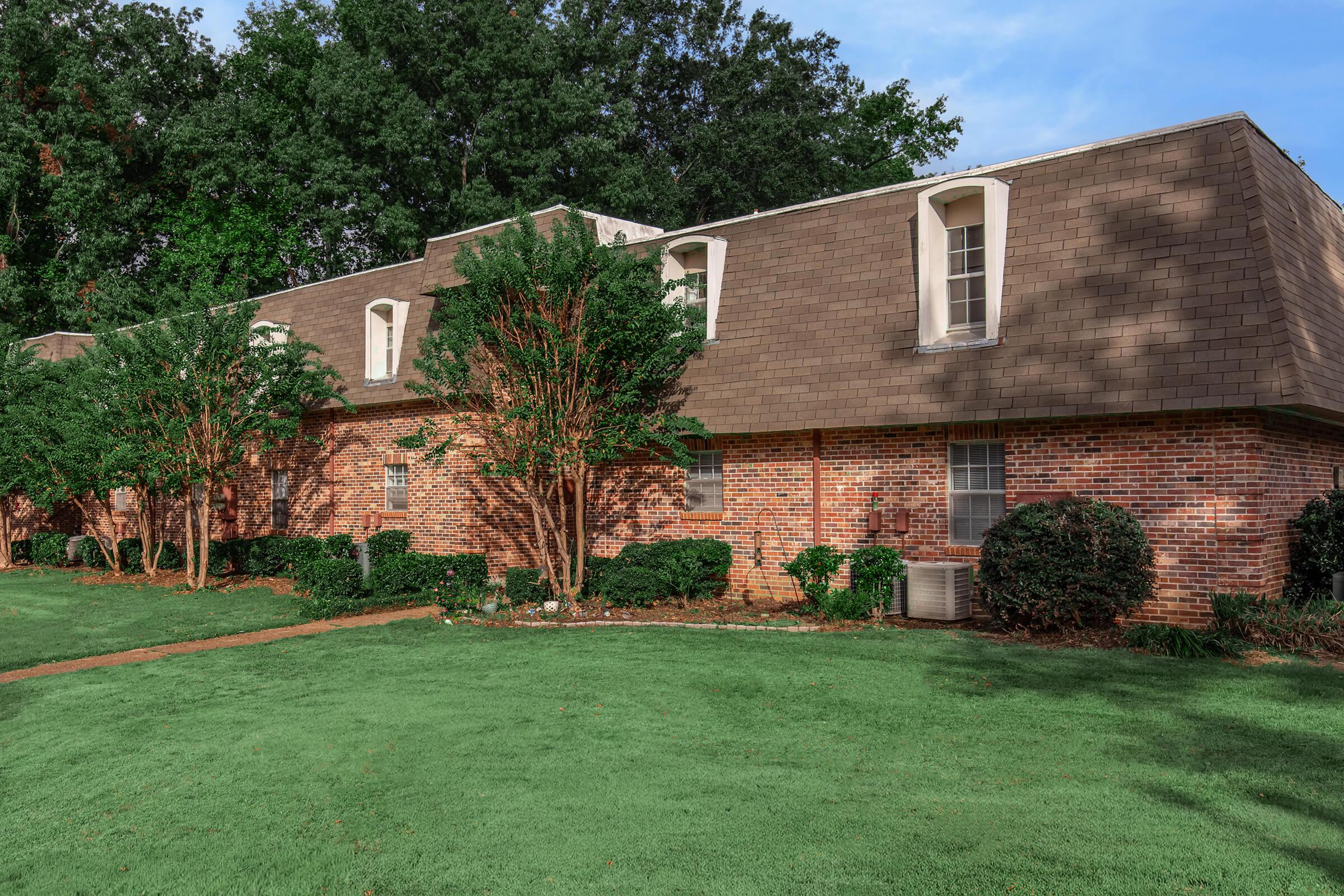 A brick building with a sloped roof, featuring several windows. The structure is surrounded by well-maintained green grass and small bushes. Trees are present in the background, contributing to a peaceful residential atmosphere.
