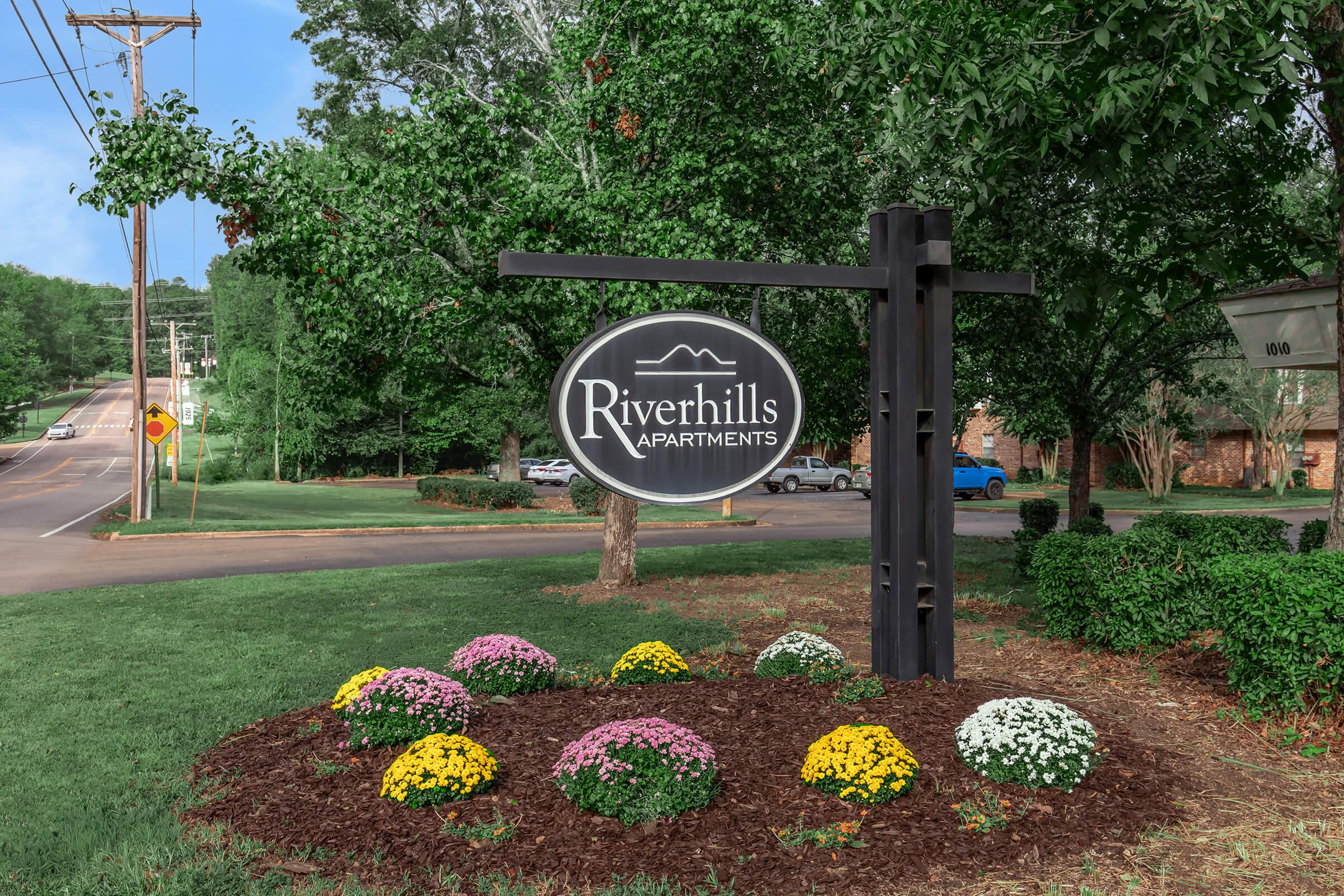 Sign for Riverhills Apartments surrounded by colorful flower beds and lush greenery. The sign features a circular design with the name "Riverhills Apartments" prominently displayed, set against a backdrop of trees and a road visible in the background.