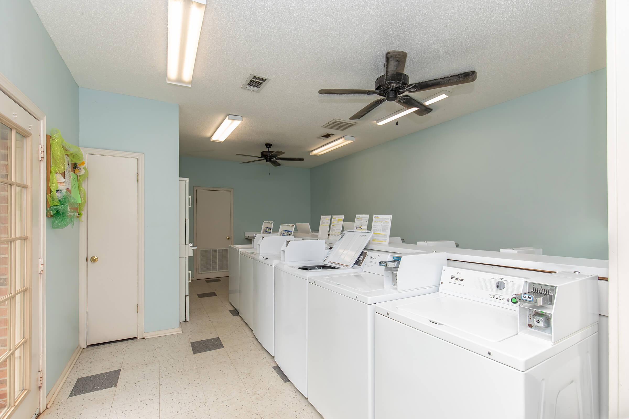 A clean laundry room featuring multiple white washing machines and dryers lined up against light blue walls. The room is well-lit with fluorescent lights and includes ceiling fans for ventilation. A door is visible in the background, along with a small area for folding clothes.