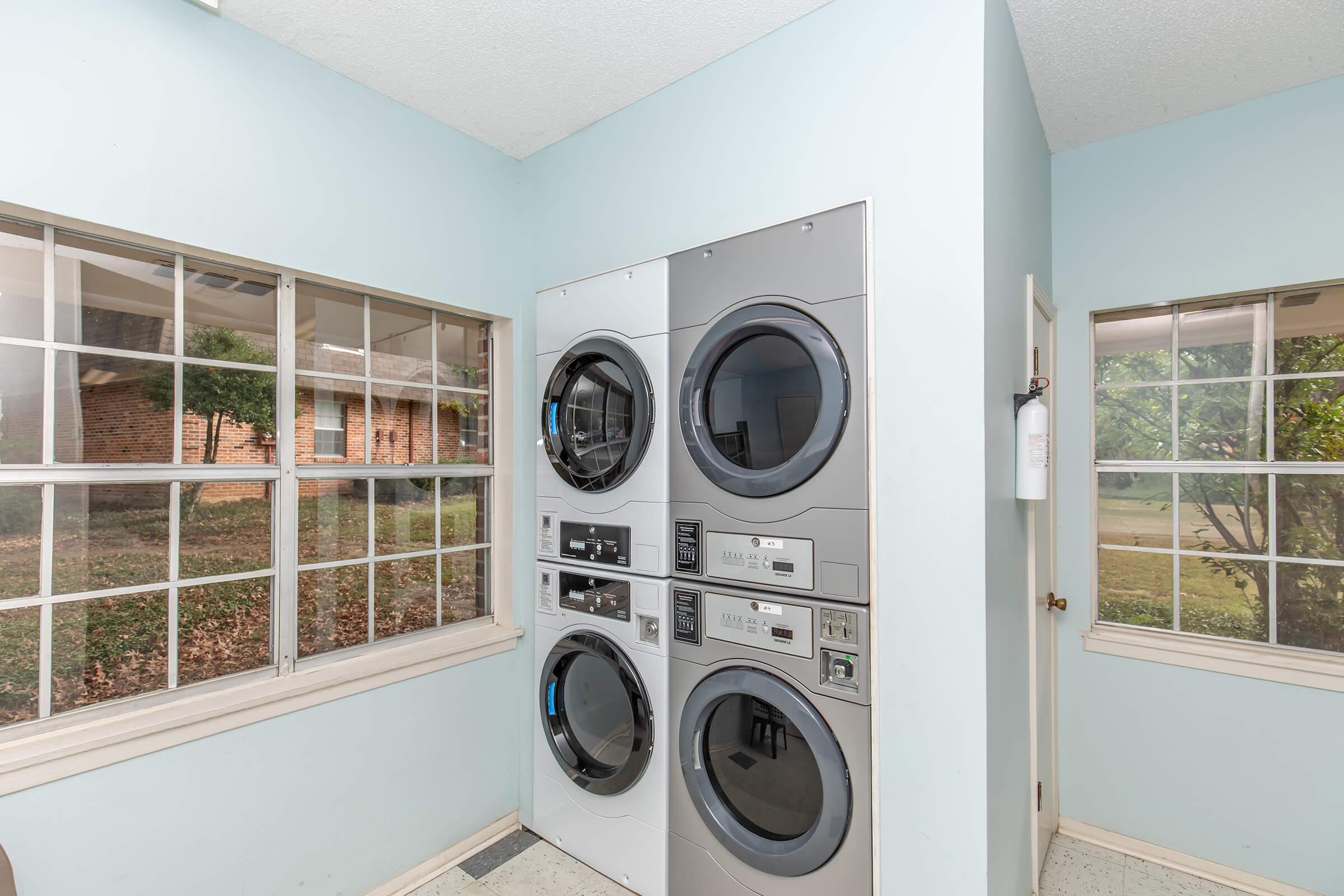 A laundry room featuring stacked washer and dryer units against a light blue wall, with large windows allowing natural light to fill the space. The view outside shows greenery and a brick building. The floor is covered with a light-colored linoleum.